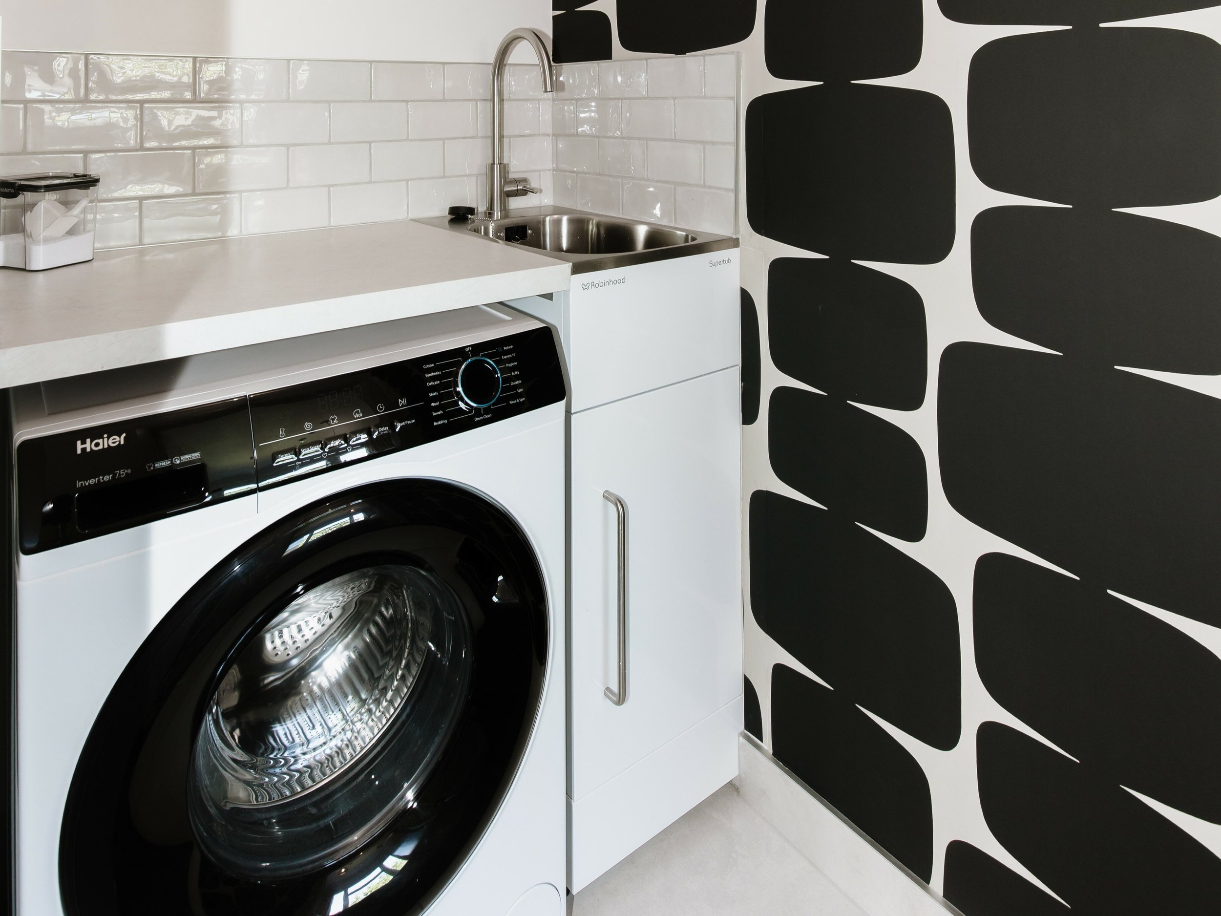 Laundry room with a front-loading Haier washing machine, a white cabinet, a small sink with a stainless steel faucet, white tiled wall behind the sink, and a black and white geometric patterned wall to the right.