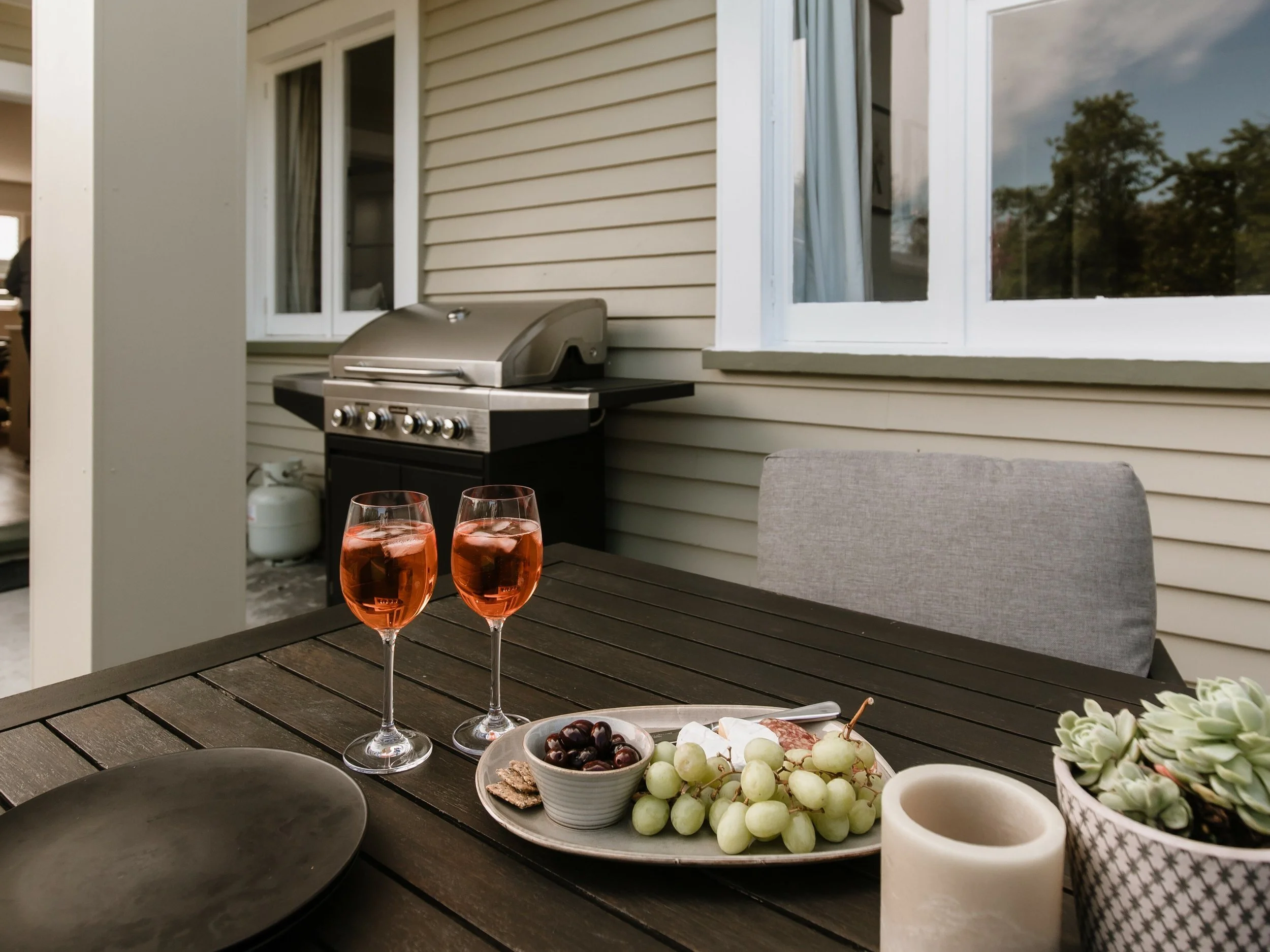 Outdoor patio table set with two glasses of rosé wine, a bowl of grapes, crackers, a candle, and a potted succulent plant, with a grill and window in the background.