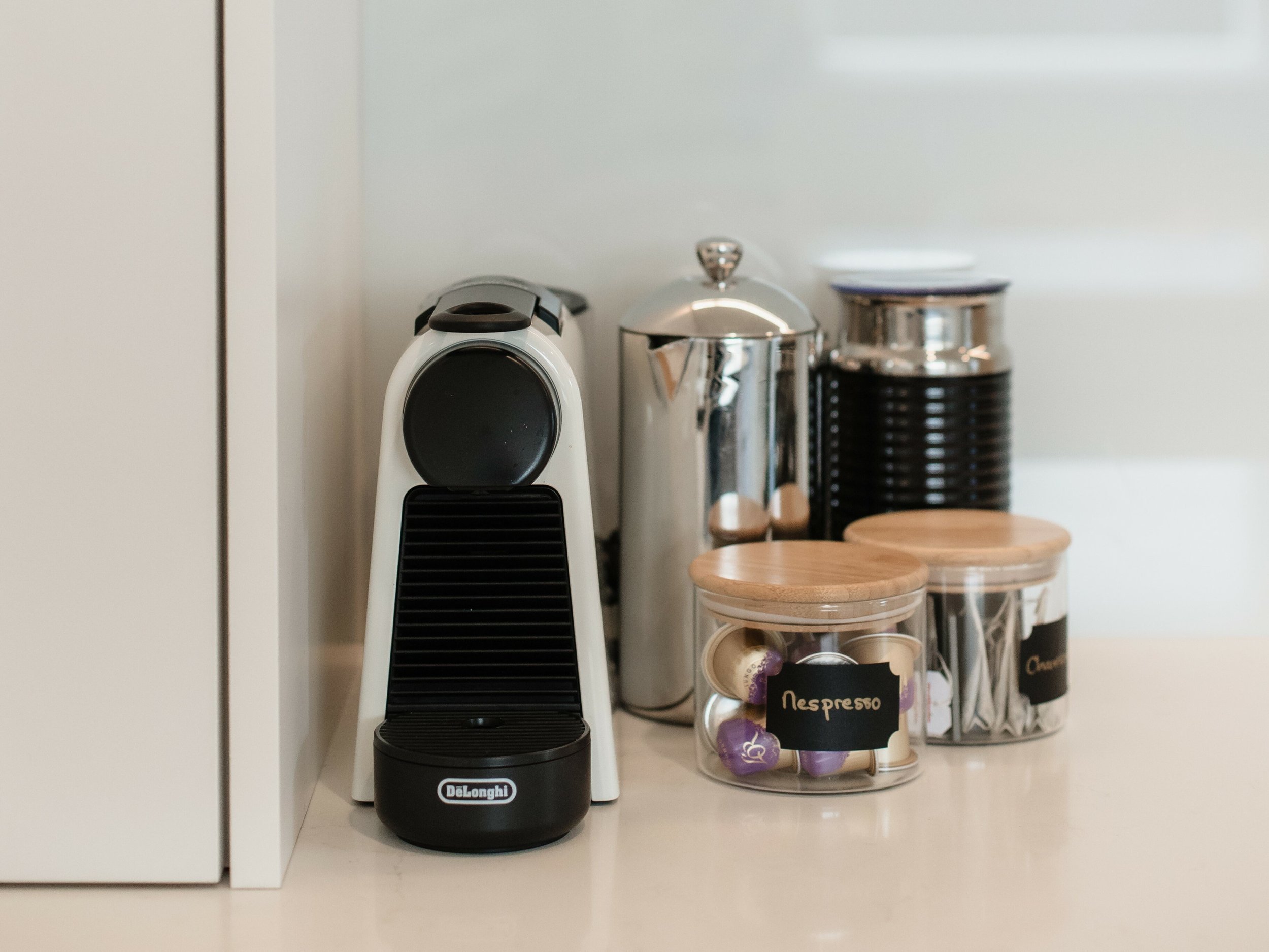 A coffee station with a DeLonghi espresso maker, a stainless steel thermal carafe, and two small glass jars labeled "Nespresso" and "Coffee" with wooden lids, containing coffee capsules and stir sticks.