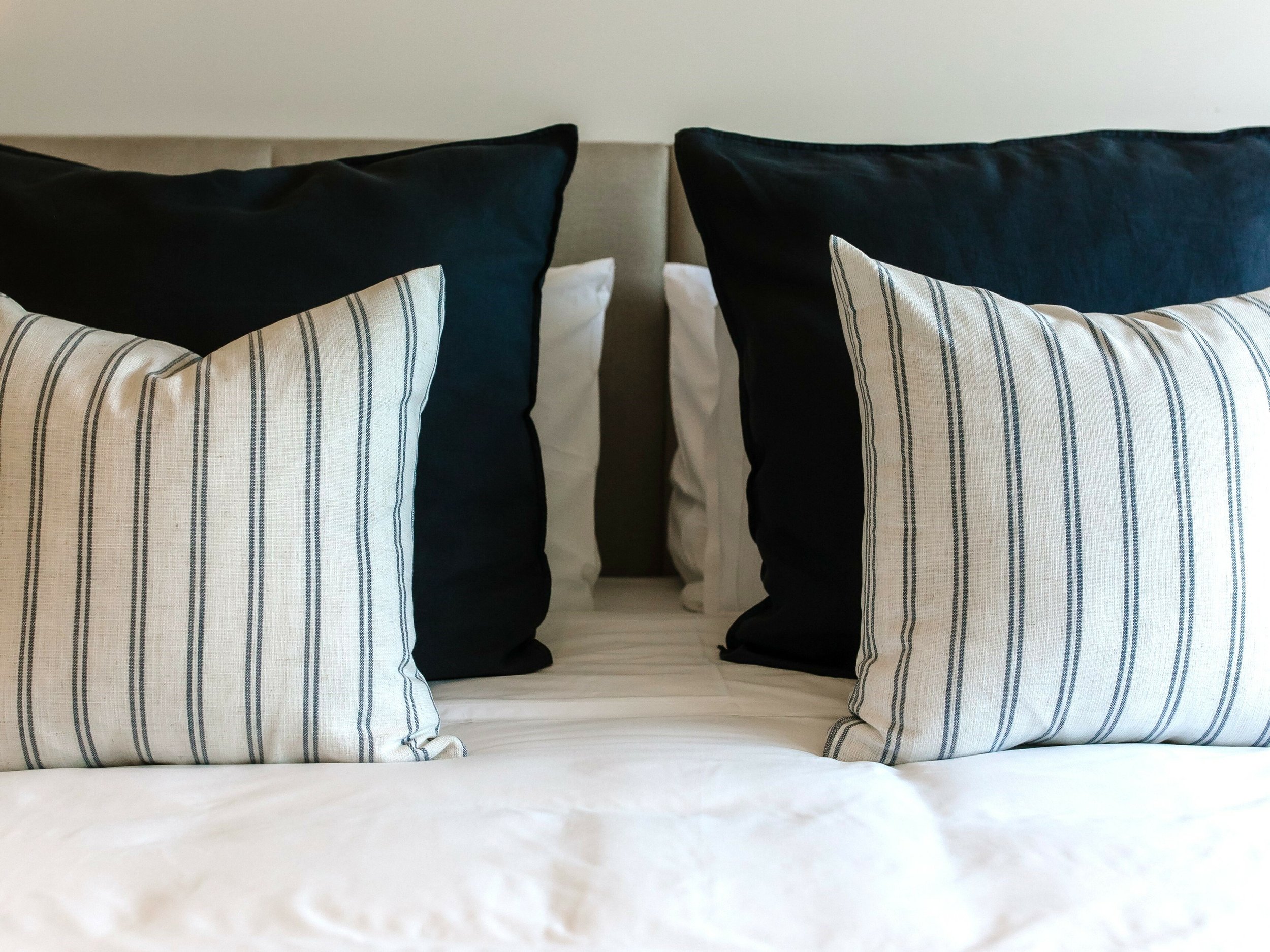 A modern bed with a beige headboard, adorned with four pillows: two larger black pillows and two smaller striped pillows with beige and black vertical lines.