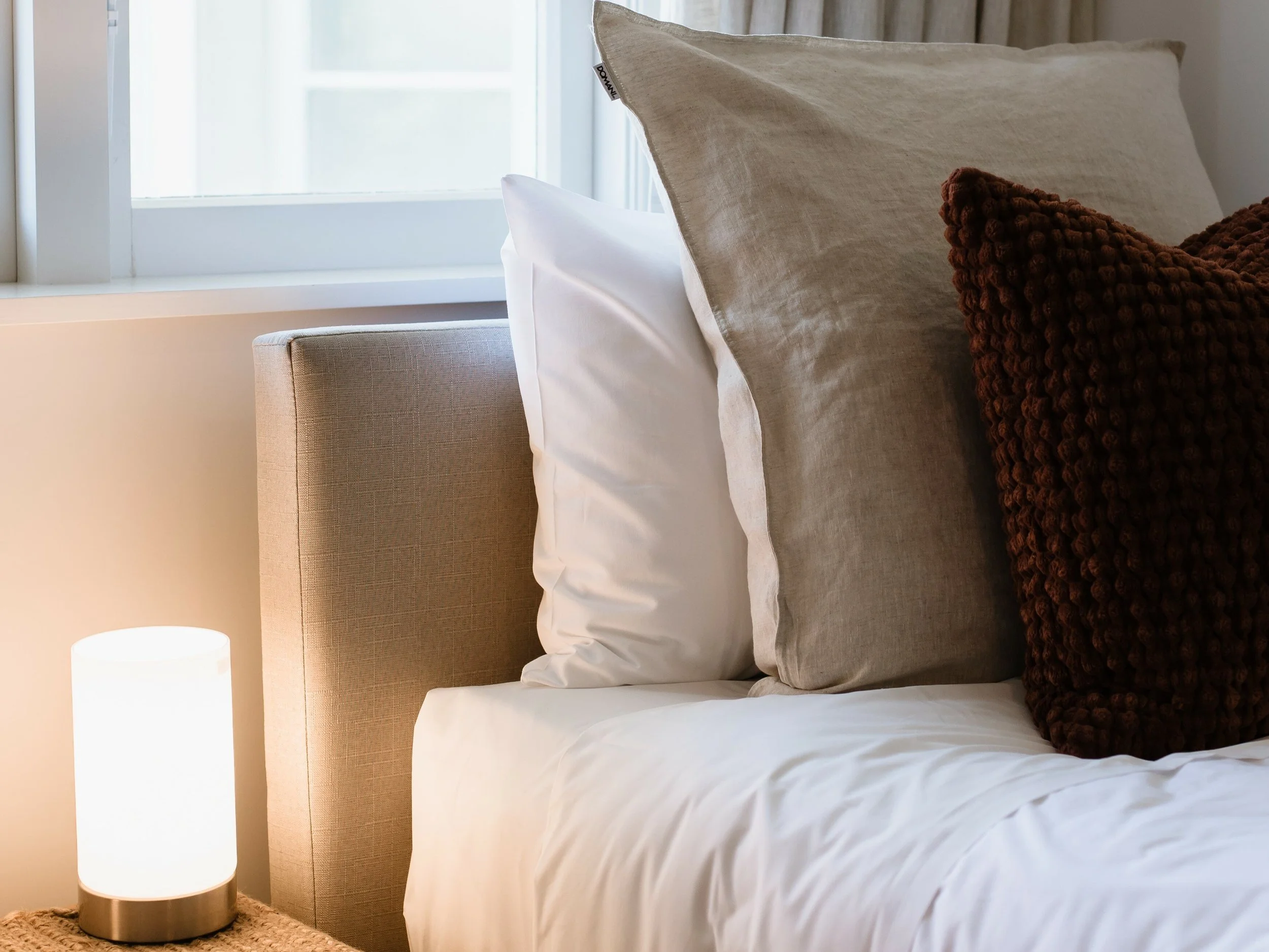 Close-up of a bed with beige headboard, white pillow, gray pillow, and textured brown pillow near a window with white curtains. Small bedside lamp on a woven surface.