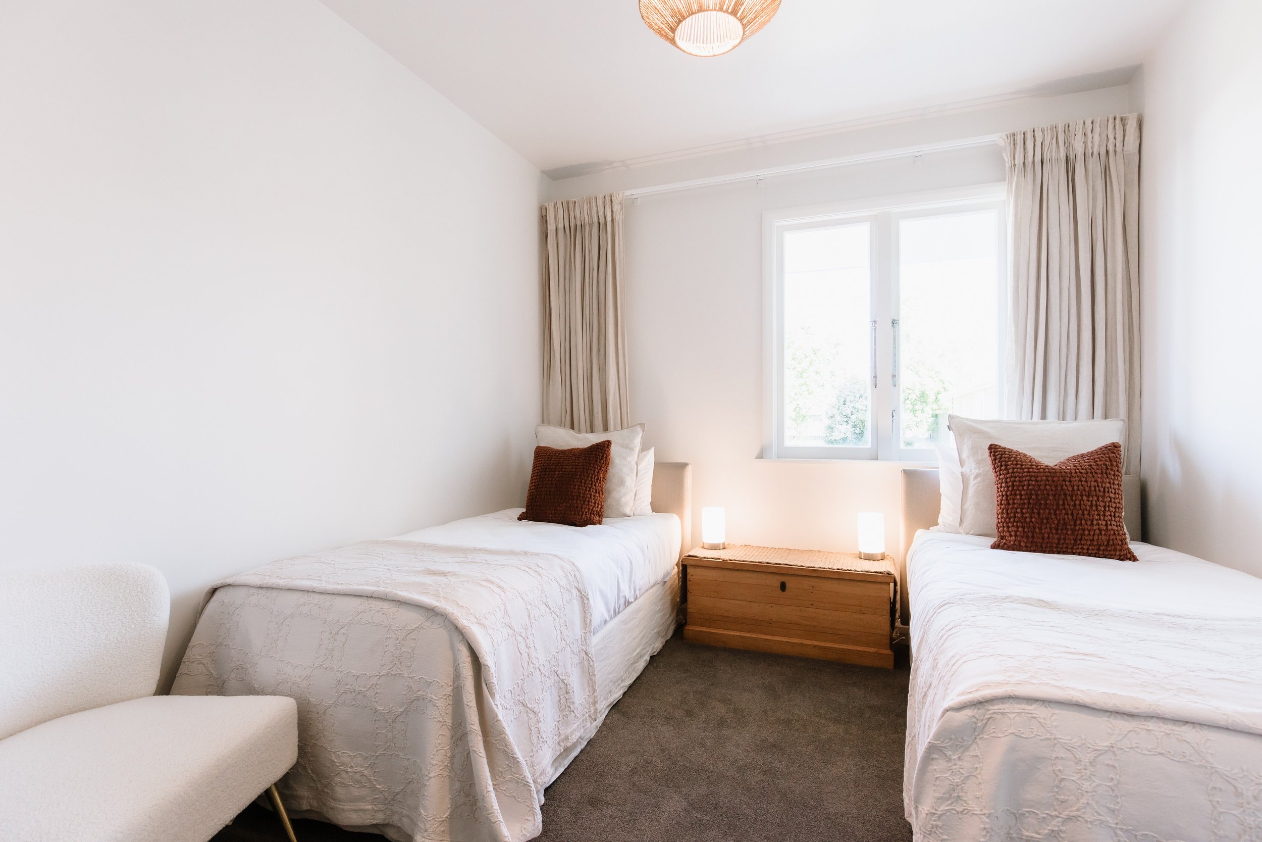 A bedroom with two twin beds, each with white bedding and a brown textured pillow. A wooden chest is placed between the beds, with two small lamps on it. There is a window with beige curtains, and a white armchair is partially visible on the left sid