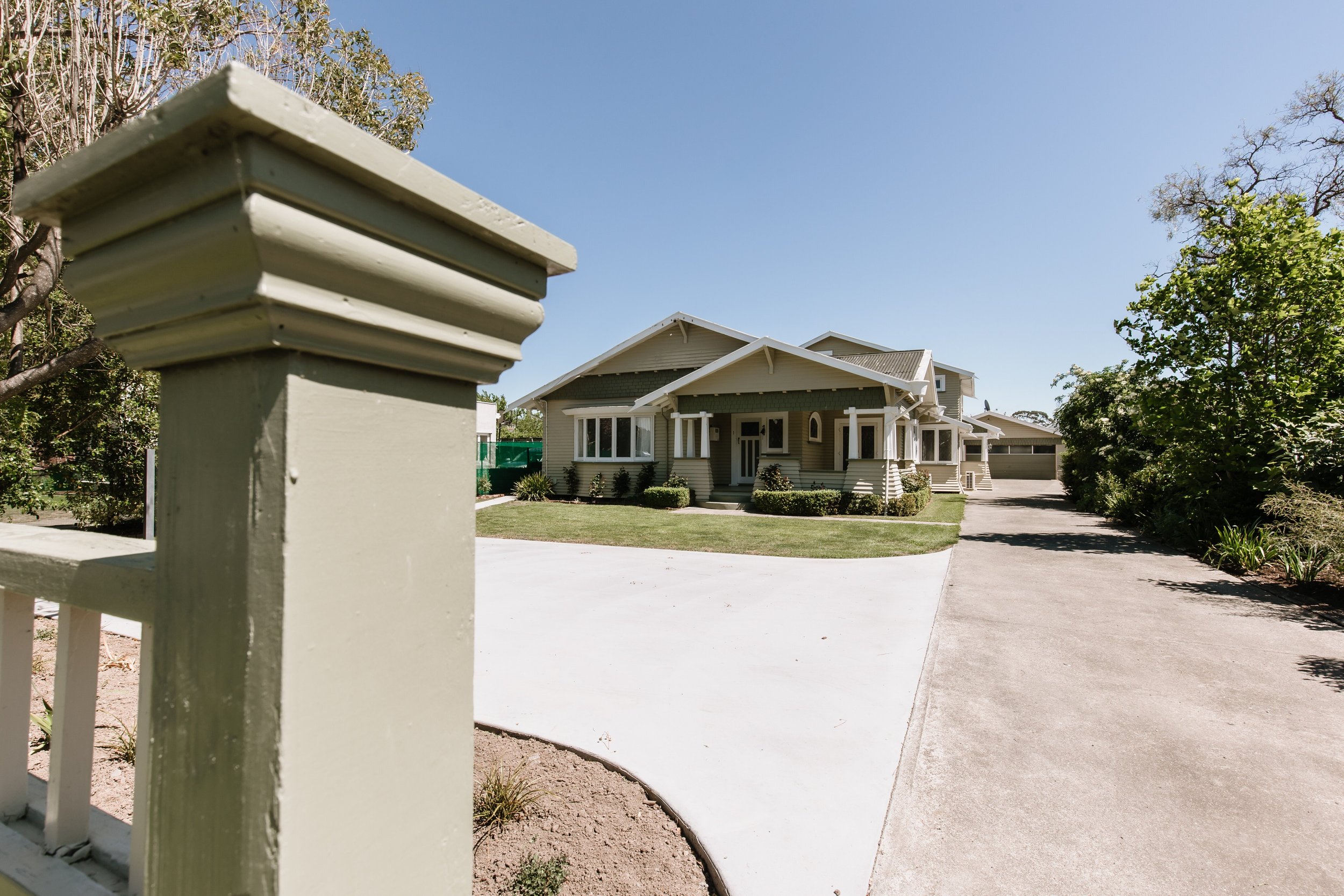 A suburban house with a well-maintained front yard, driveway, and a beige picket fence post in the foreground on a sunny day.