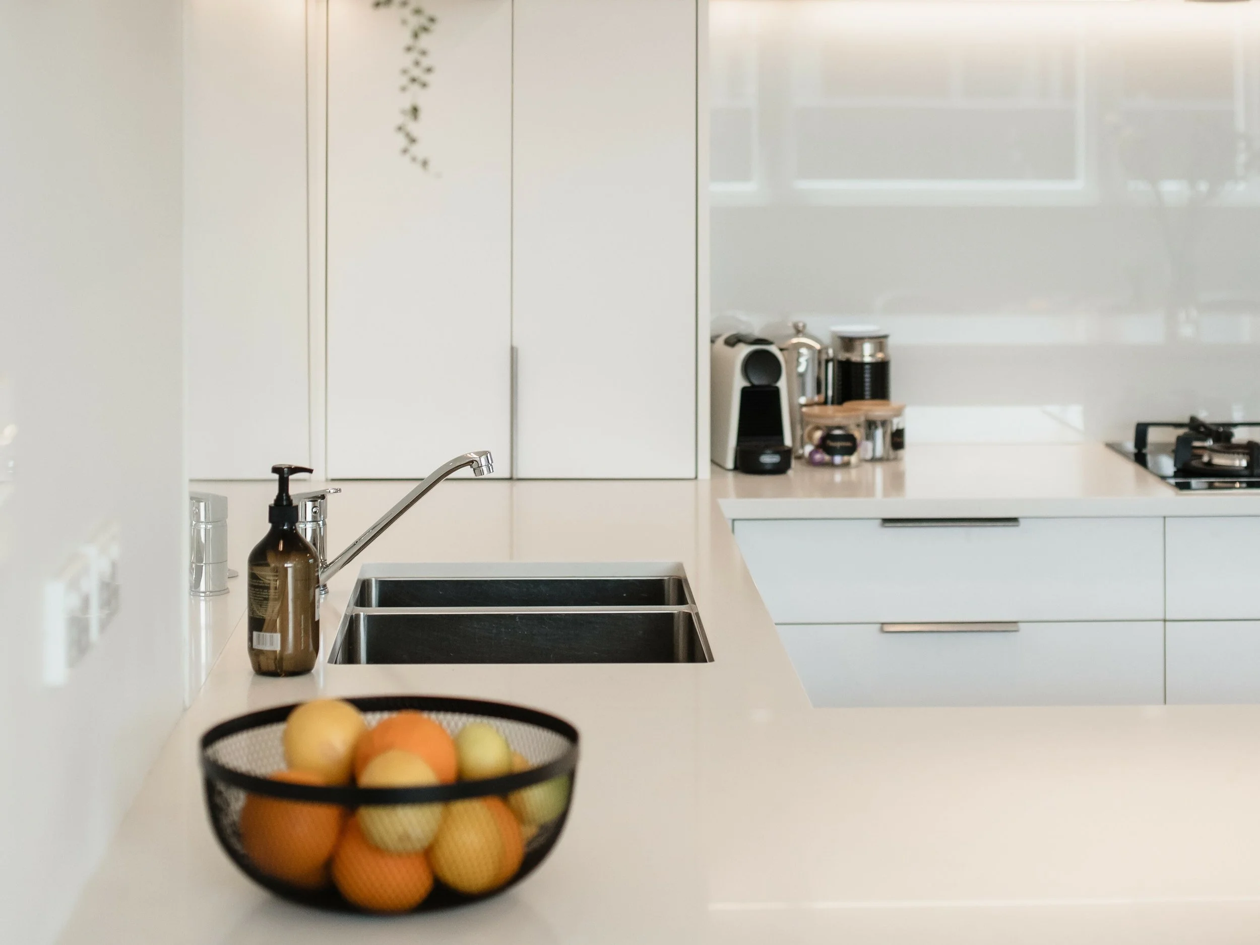 Modern kitchen with white cabinets, a black fruit basket filled with oranges and lemons, and various small appliances on the countertop.