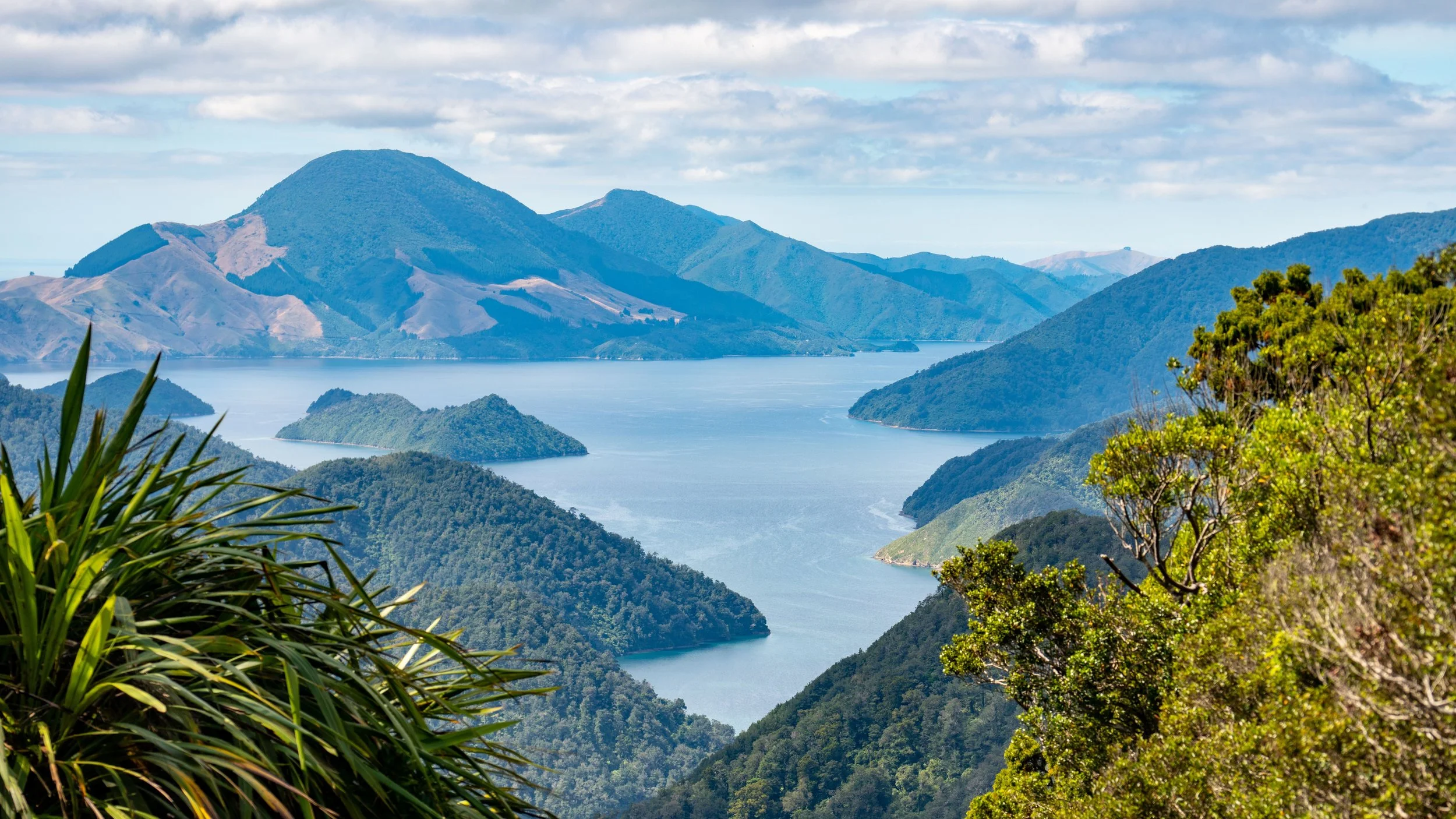 A scenic view of a lake surrounded by lush green mountains and hills with partly cloudy sky.