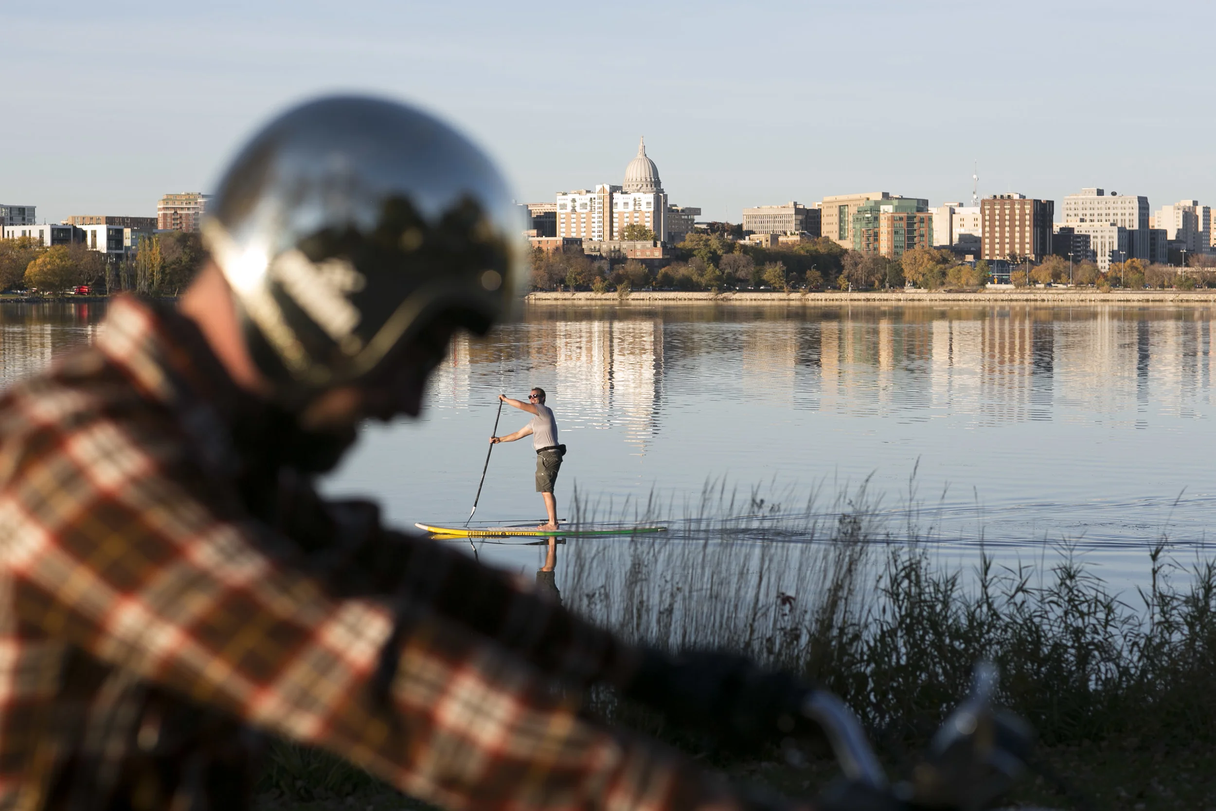 Chris Brand prepares to ride his 1974 Harley Davidson as a paddle-boarder drifts past in Monona Bay. Brand restores vintage motorcycles with his brother in their home welding shop on the east side of Madison.