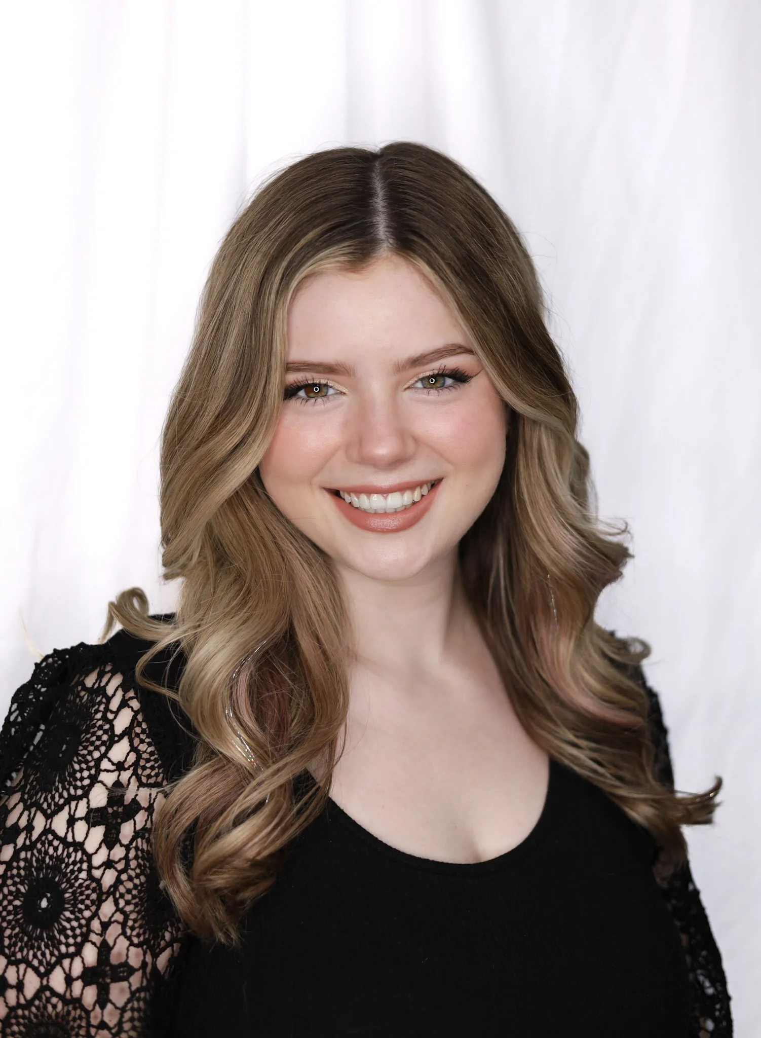 Portrait of a young woman with long wavy light brown hair, smiling, wearing a black top with lace sleeves, standing against a white background.