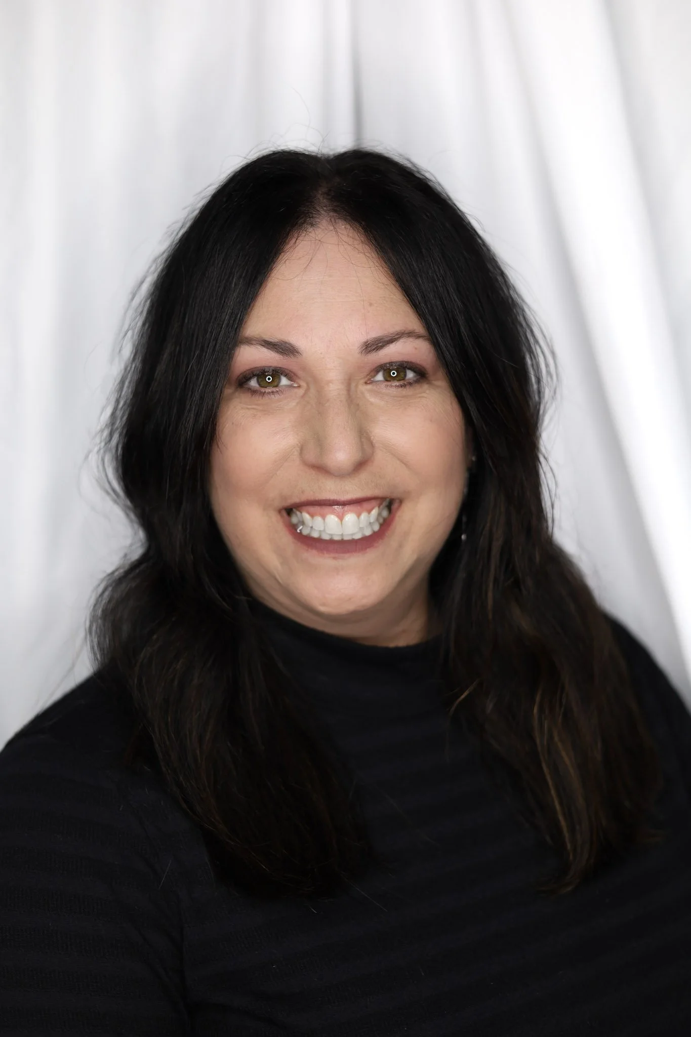 A woman with shoulder-length dark hair and light skin, smiling in front of a silver curtain background, wearing a black top.