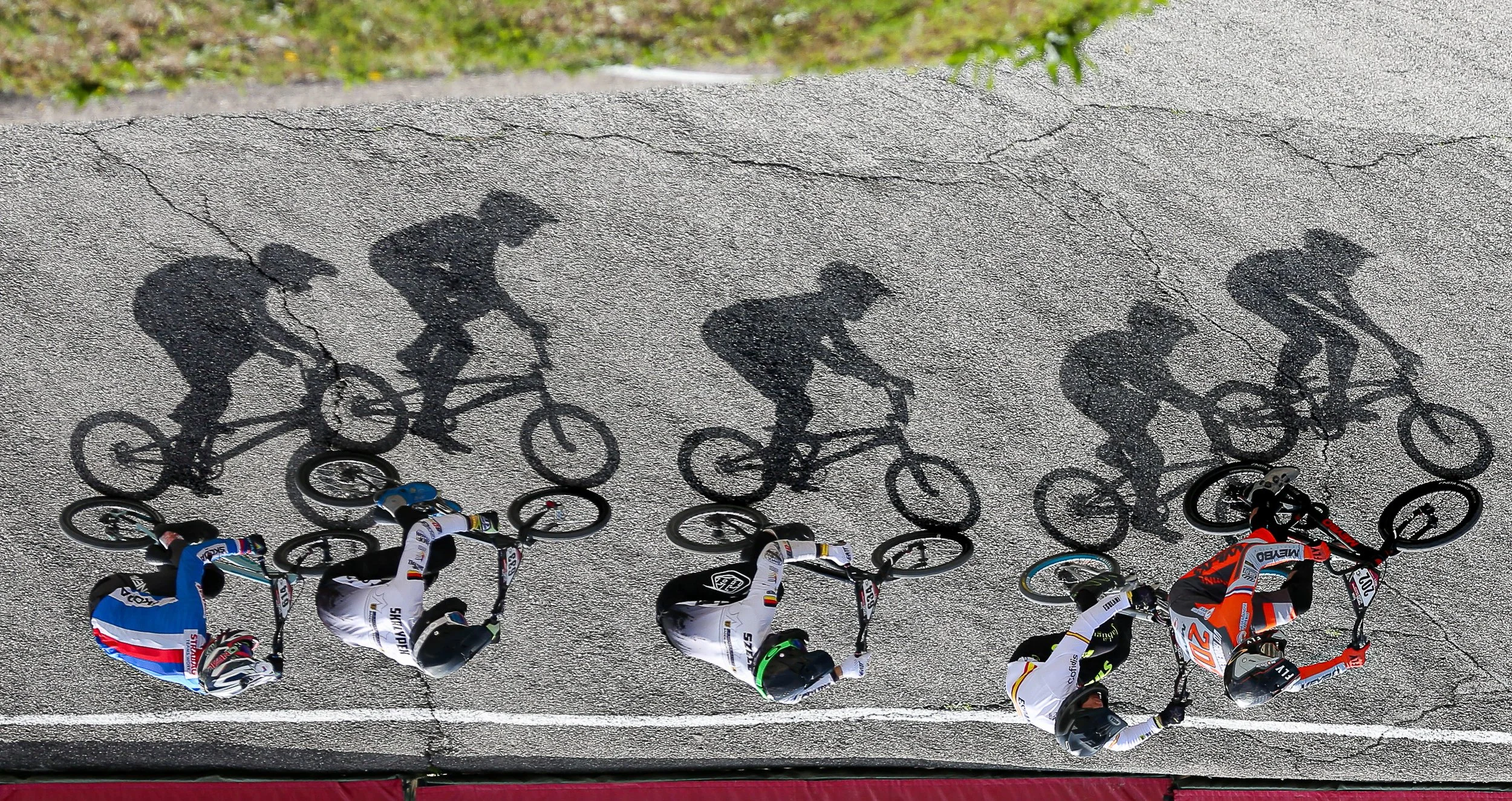 Five BMX riders race across sunlit asphalt, their elongated shadows forming a second line above them in Mickael Chavet’s award-winning photograph The Shadow Line