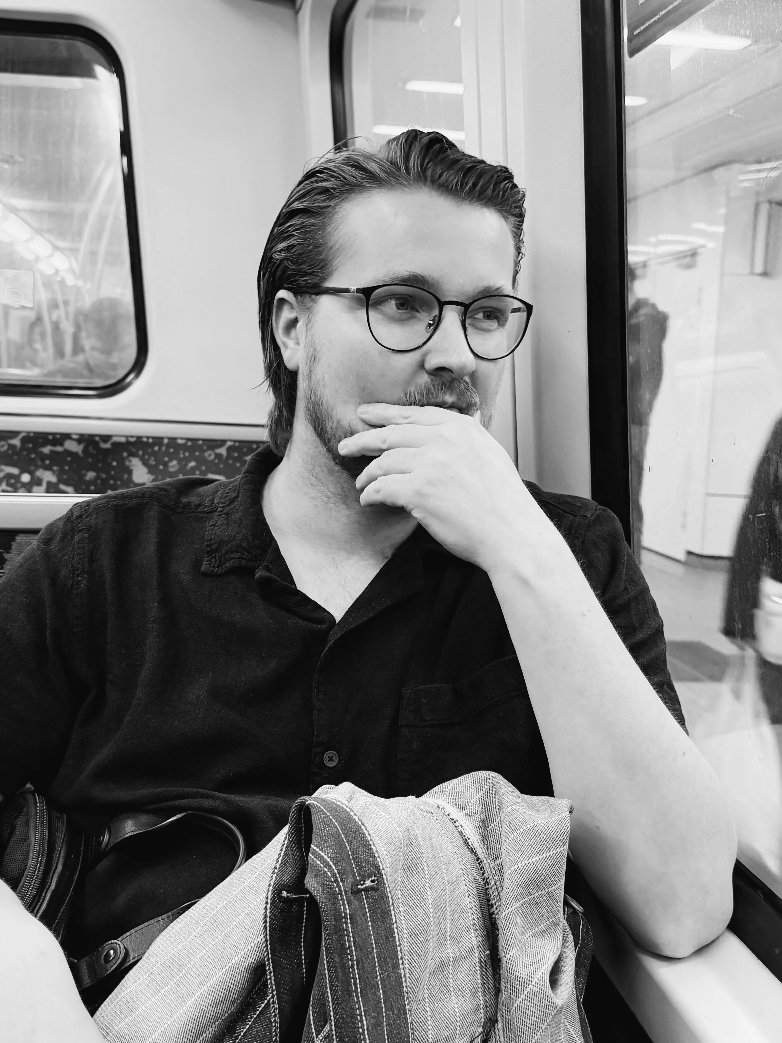 A young man with glasses and a beard, sitting on a train, looking out the window and resting his chin on his hand.