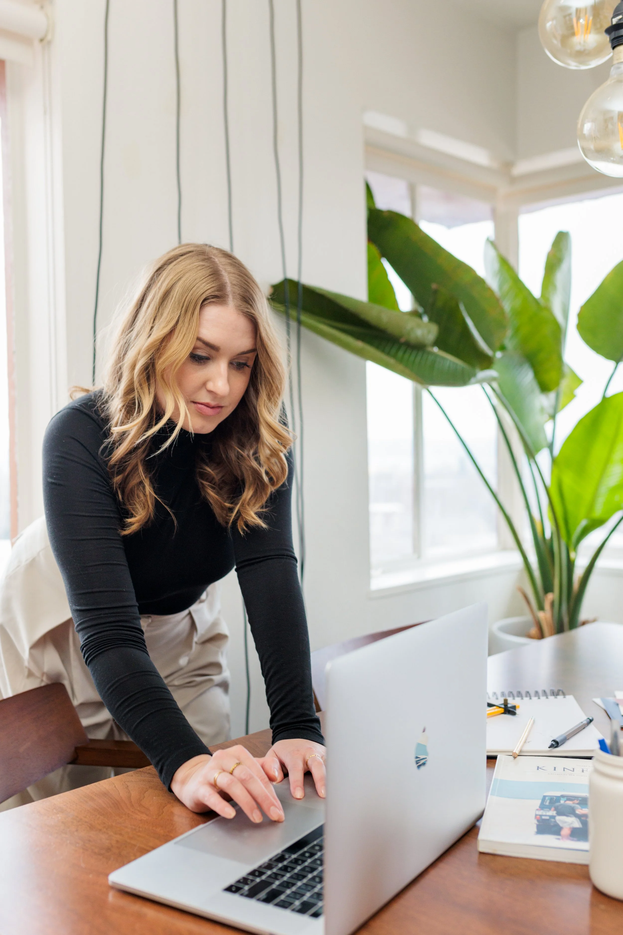 A woman in a black long sleeve shirt and beige pants leaning over a wooden table, working on a silver laptop. There are papers, pens, and a magazine on the table, with large green plants and windows in the background.