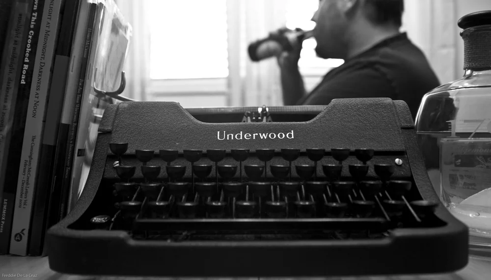 An Underwood typewriter in the foreground; Hosho McCreesh drinks from a bottle by a bright window behind it, black and white