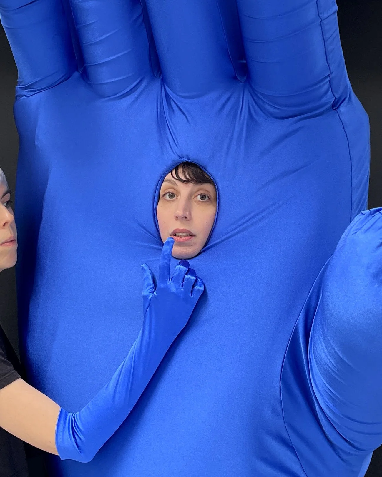 Still from a performance by artist Colleen Asper. A face peeking through the palm of a giant blue inflatable hand while a gloved finger touches their lips, against a black backdrop.