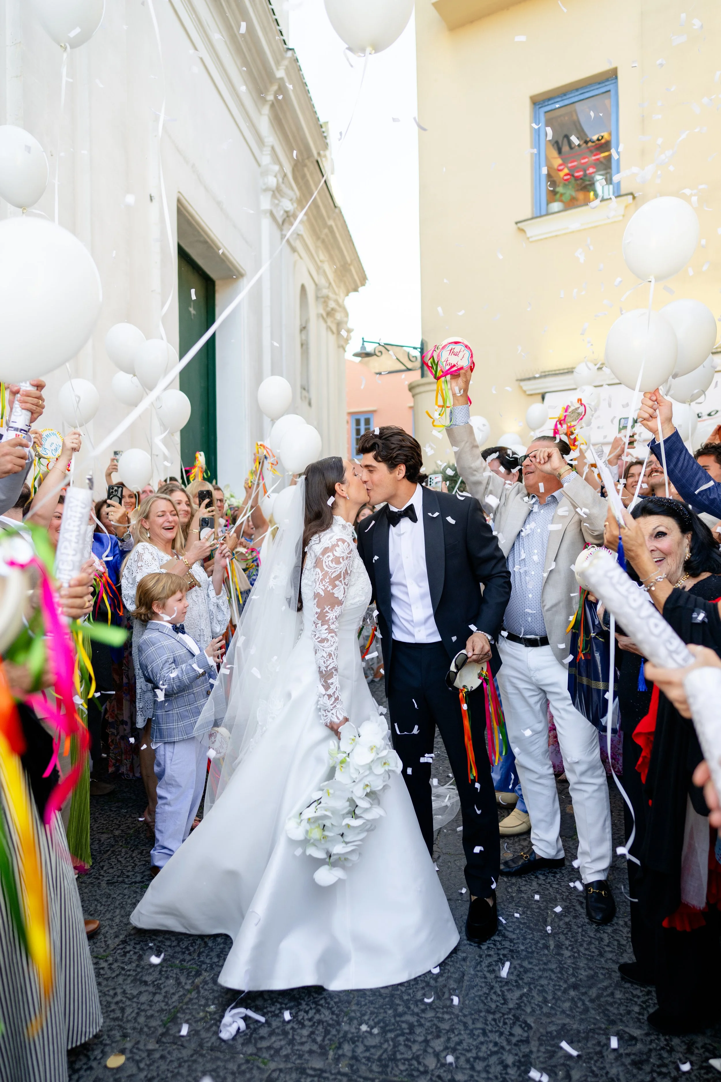 Stella Blue bride Rachel and groom Franco kiss outside Chiesa di Santo Stefano in Capri, Italy, surrounded by wedding guests, white balloons, and falling confetti.
