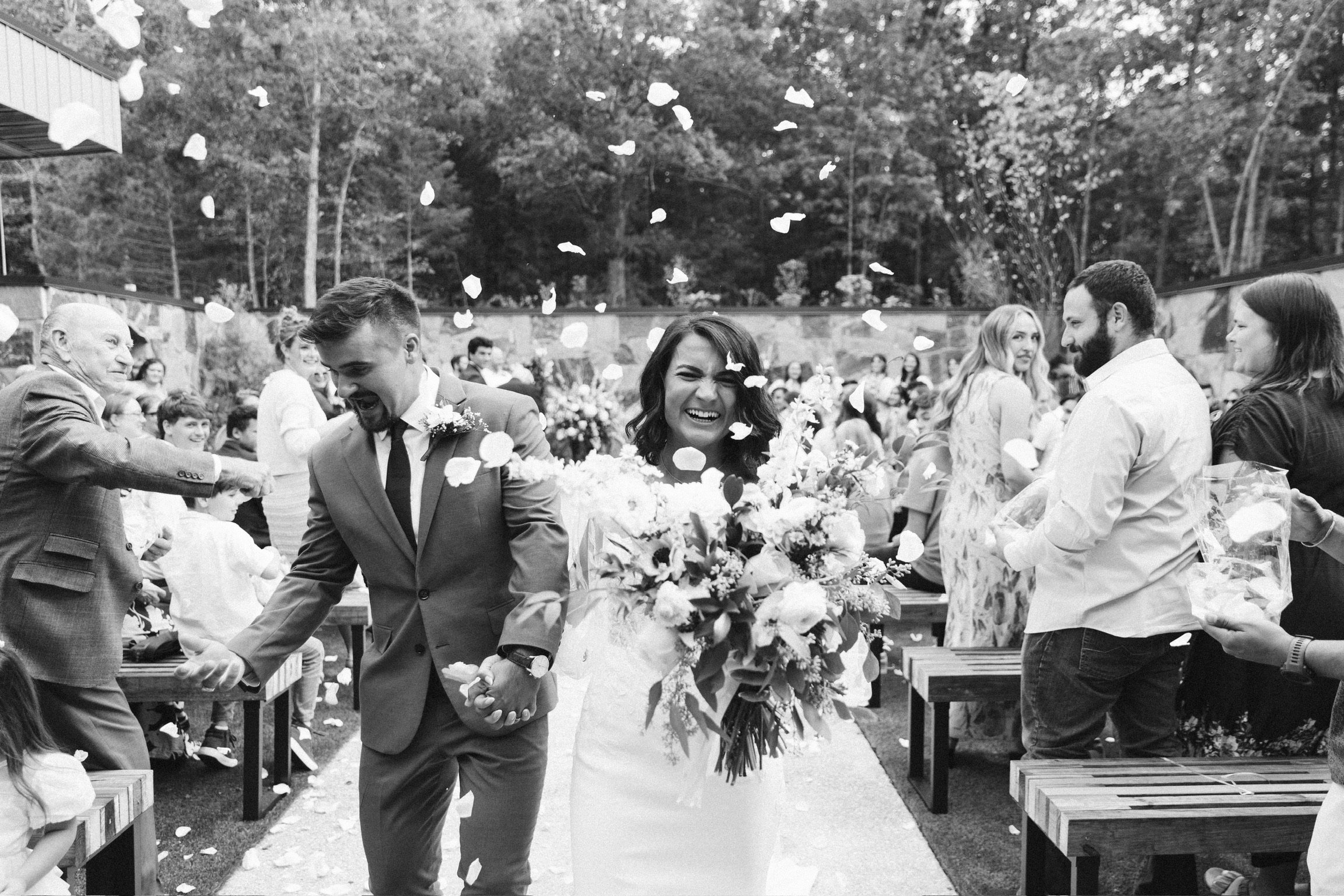 Stella Blue Design bride and groom Lana and Alex walk hand in hand down the aisle after their wedding ceremony as guests toss flower petals around them. Lana carries a large floral bouquet.