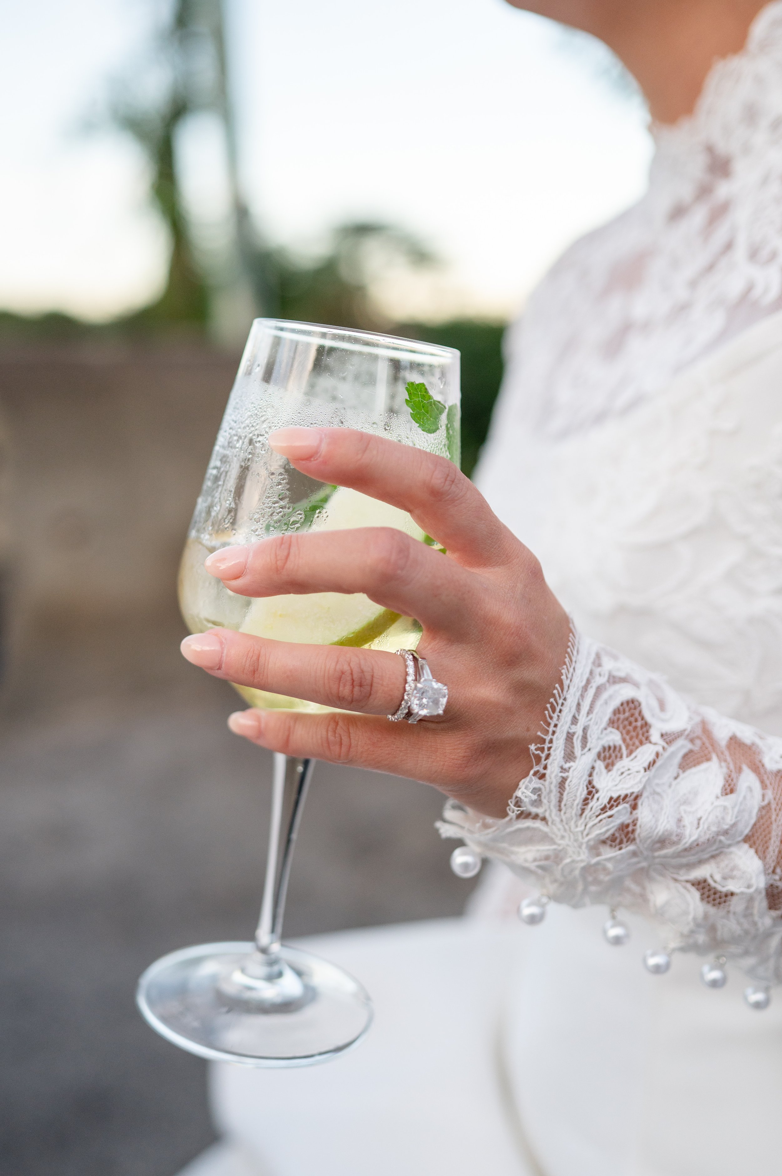 Stella Blue bride Rachel holds a celebratory mojito, showcasing her 4-carat elongated cushion-cut diamond engagement ring with tapered baguette side stones and round diamond wedding band. She wears a lace Monique Lhuillier wedding gown.