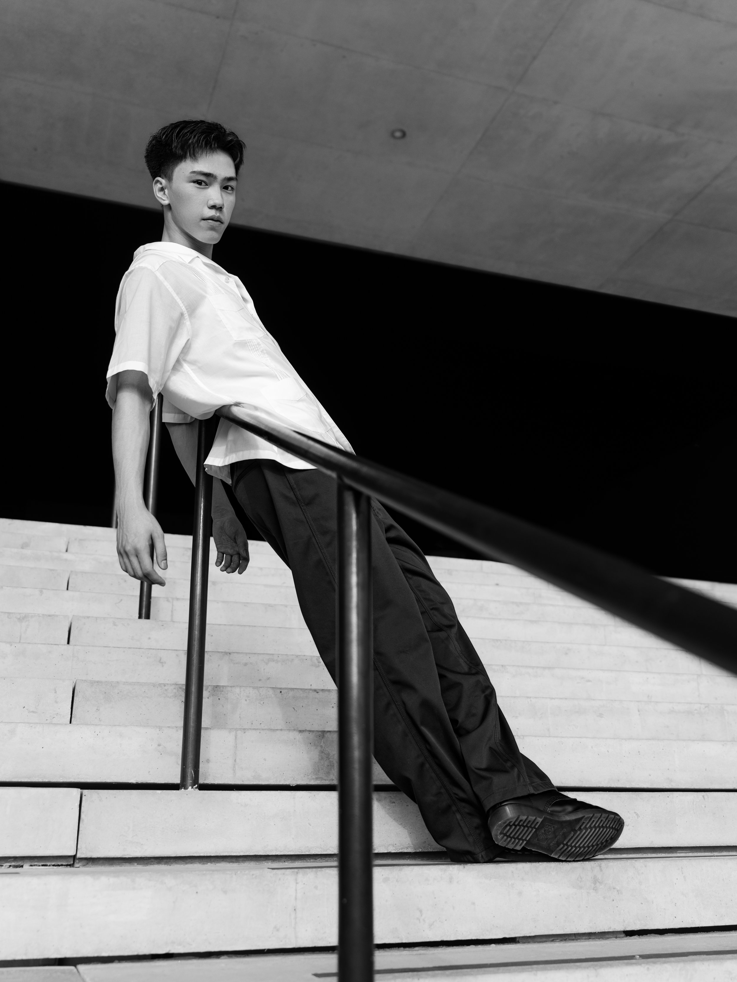 A young man with dark hair leaning against a staircase handrail, looking at the camera, in a modern indoor space with a concrete ceiling.