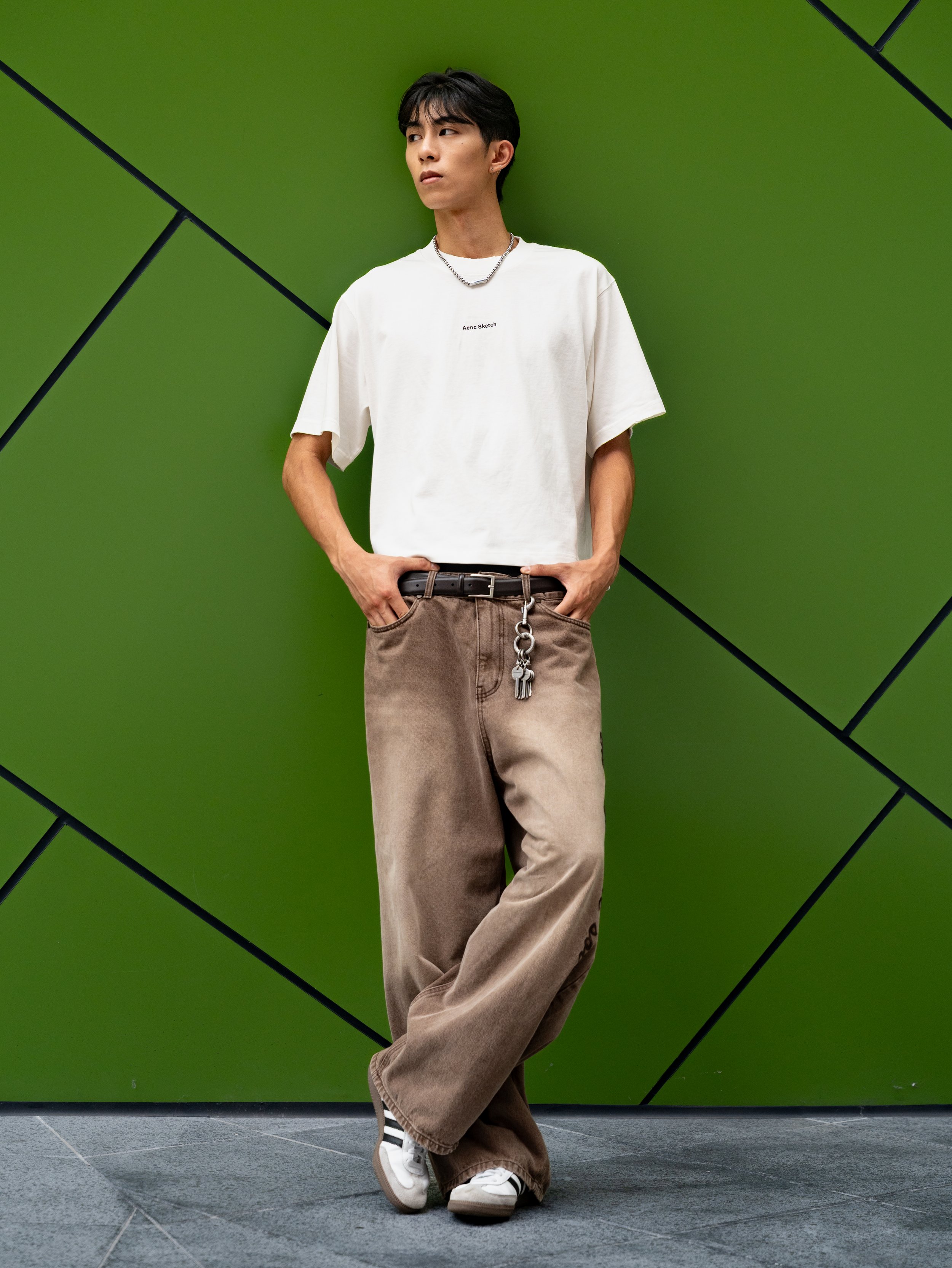 A young man stands relaxed against a vibrant green wall with black diagonal lines, wearing a white oversized T-shirt, beige baggy pants, and white sneakers with black stripes. He accessorizes with layered necklaces and has dark hair styled in a casua