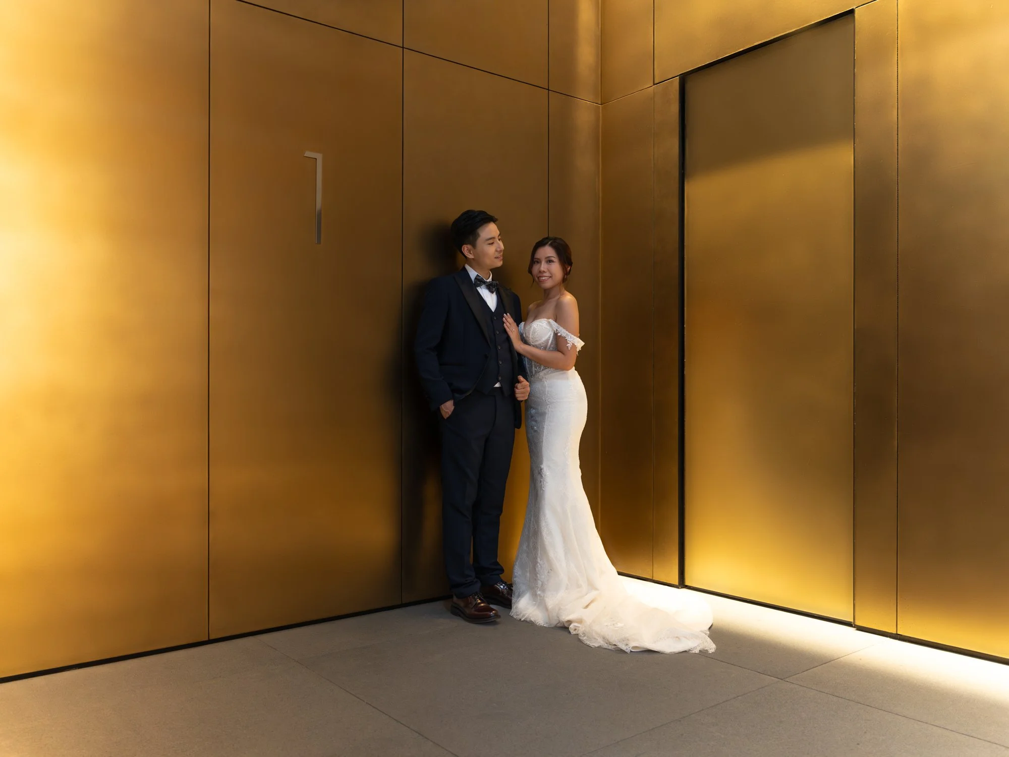 A newlywed couple stands in an elevator lobby with gold-colored walls. The groom wears a navy suit and bow tie, and the bride wears a white off-the-shoulder wedding gown. They are smiling and looking at each other.