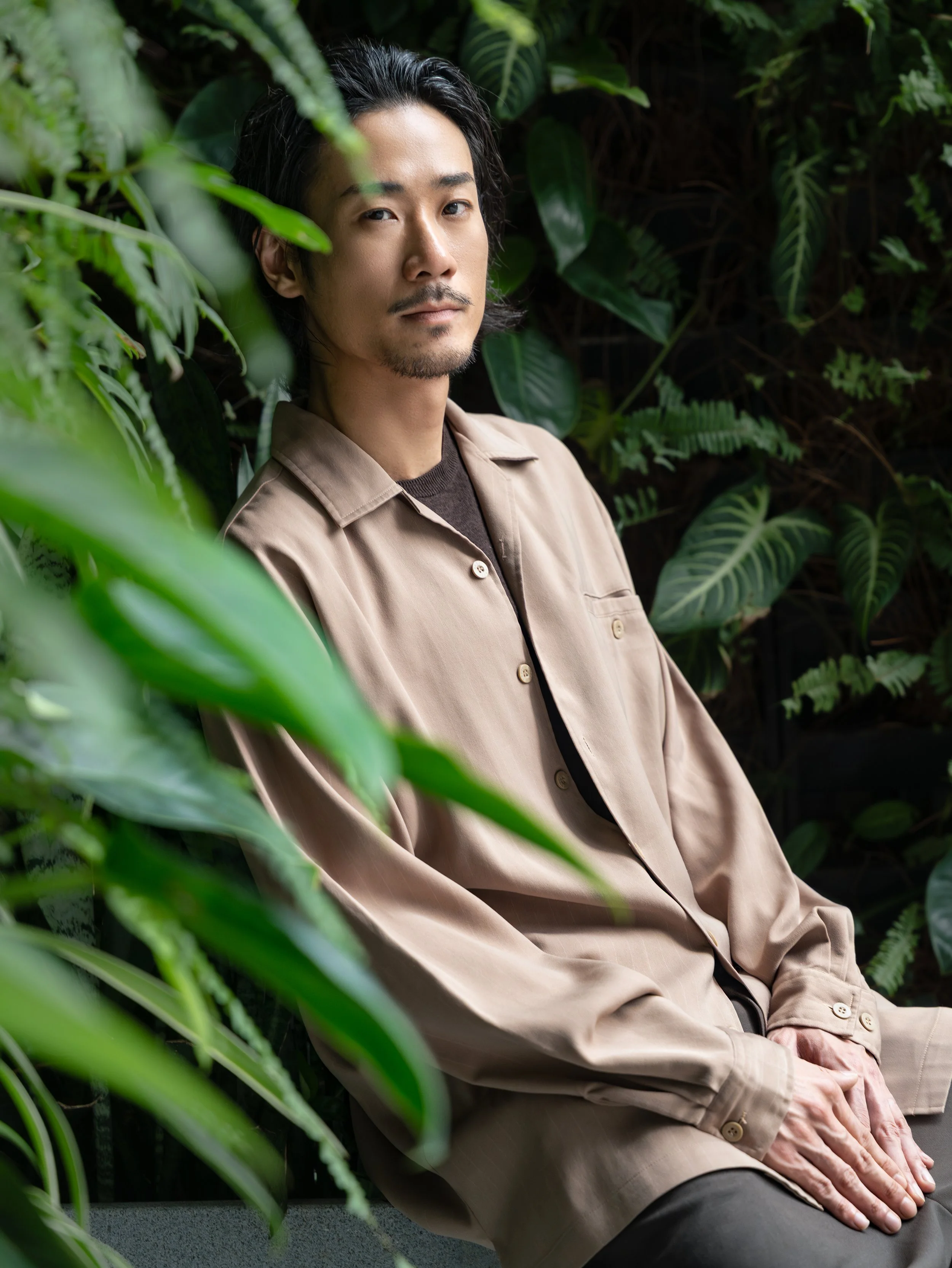 A man with dark hair, a slight mustache, and a goatee, wearing a light beige coat and black shirt, sitting among lush green plants with a contemplative expression.