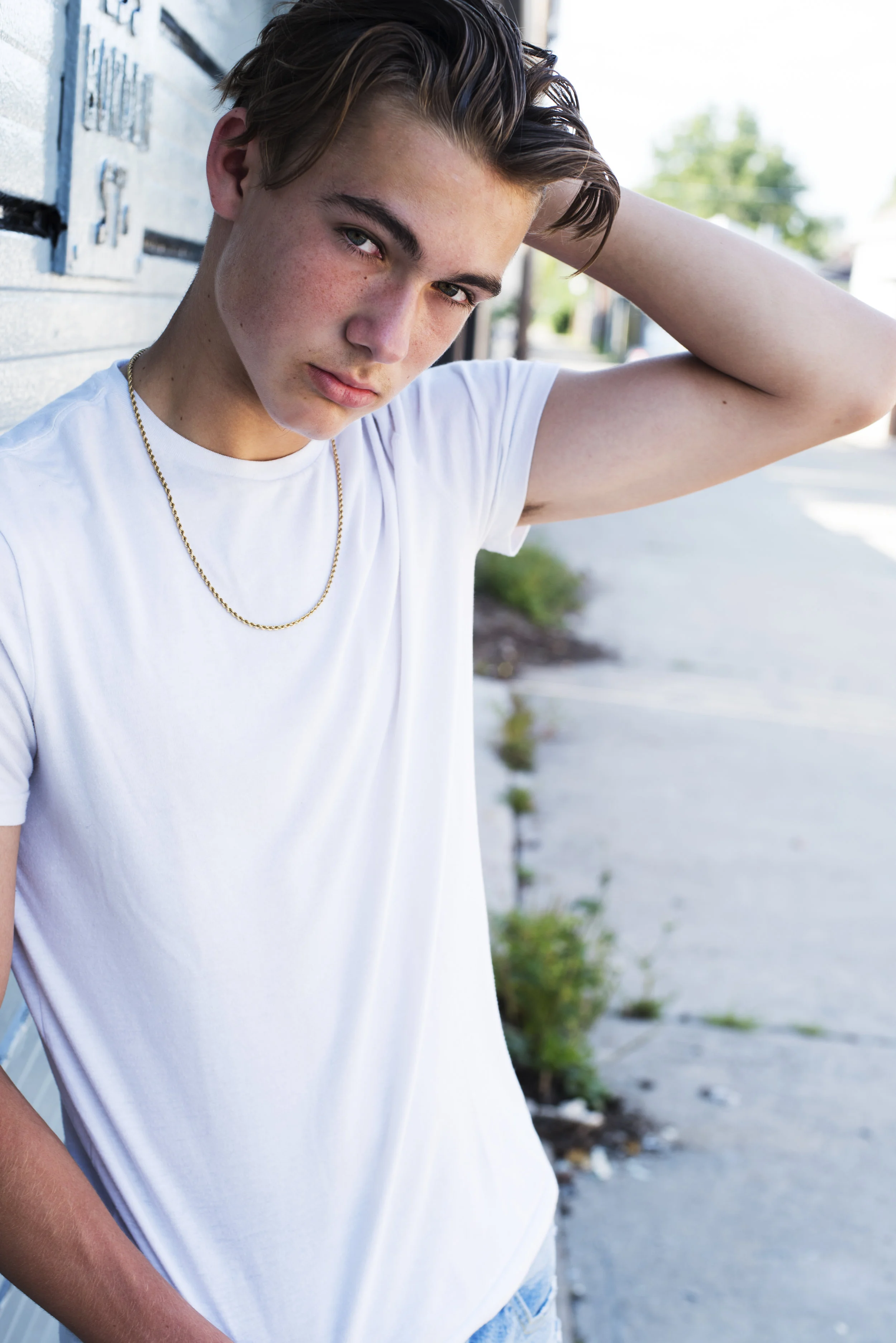 A teenage boy with light skin, freckles, and blue eyes standing outdoors next to a light gray wall, looking at the camera with a serious expression, wearing a white t-shirt and a gold chain necklace, with one arm raised and hand resting on his head, 