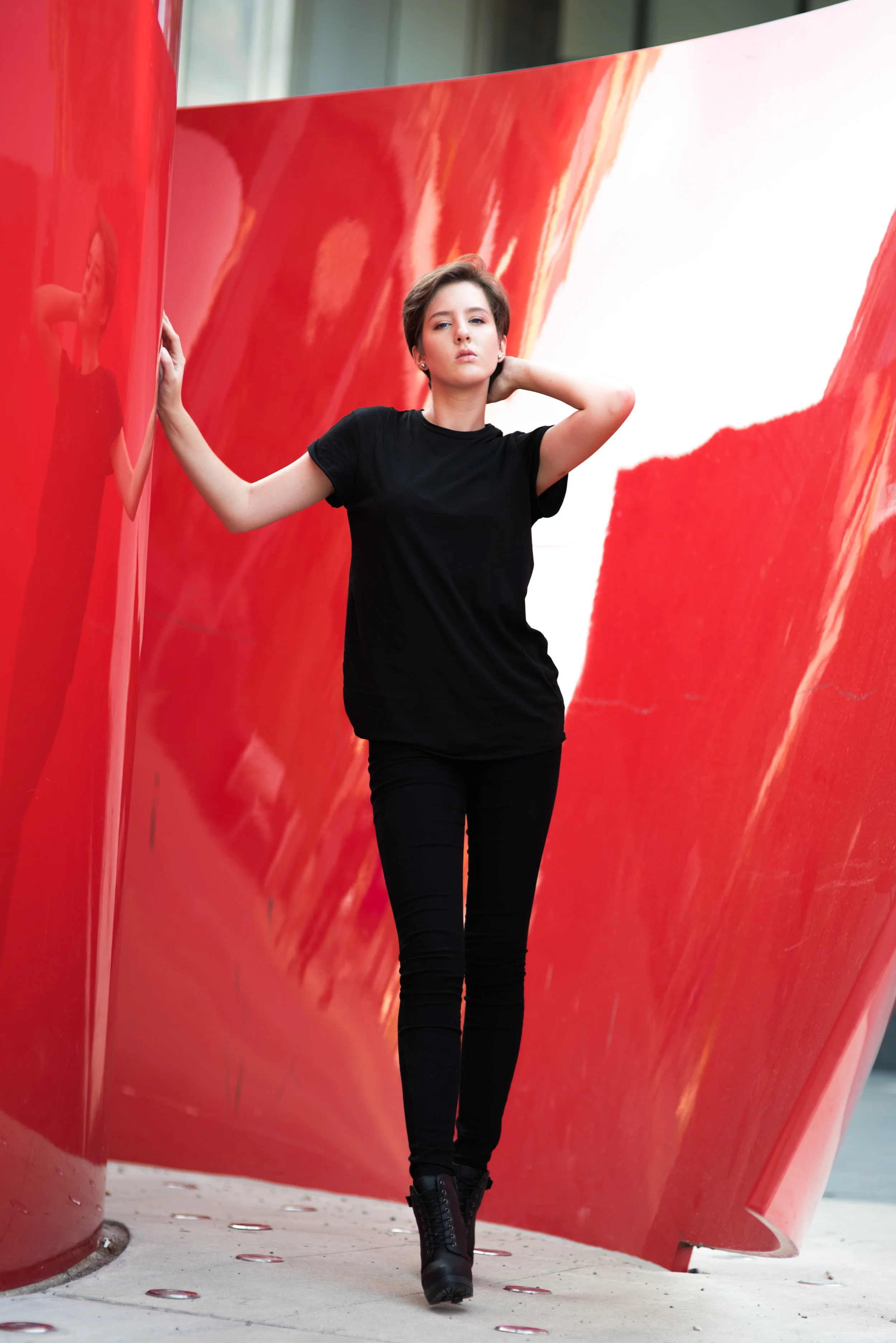 A woman with short brown hair wearing a black T-shirt black pants and black combat boots walking between large red modern sculptures outdoors.