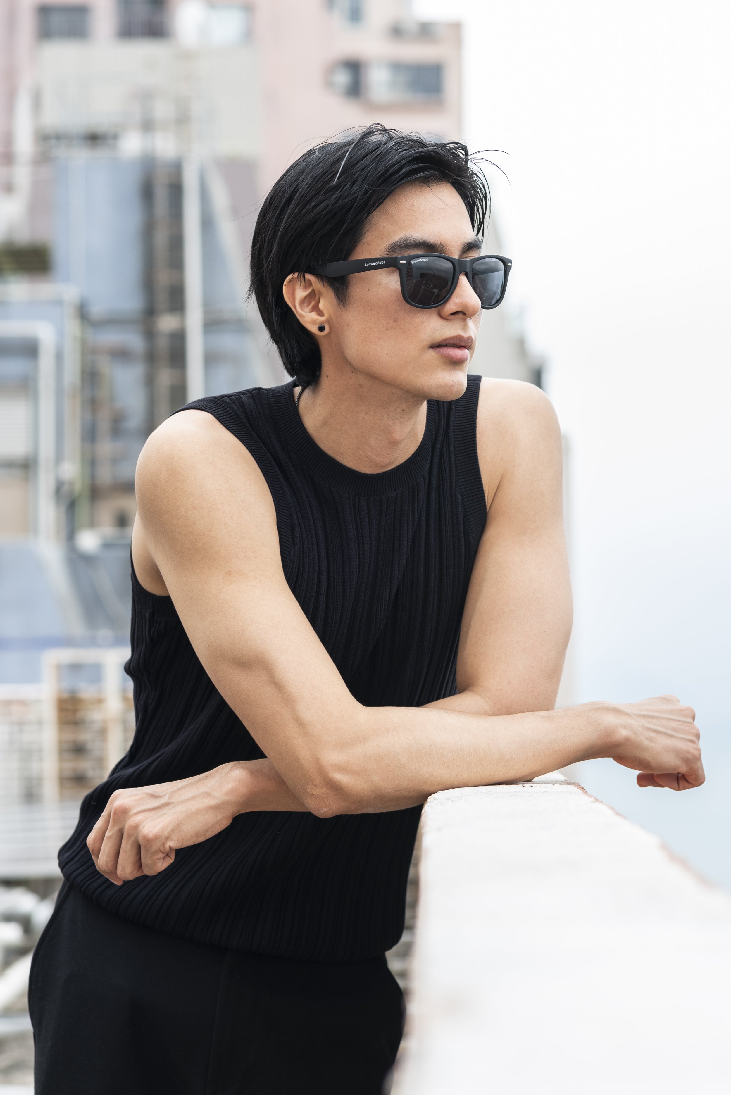 A woman with short black hair wearing black sunglasses and a sleeveless black top, leaning on a white railing on a balcony with city buildings in the background.