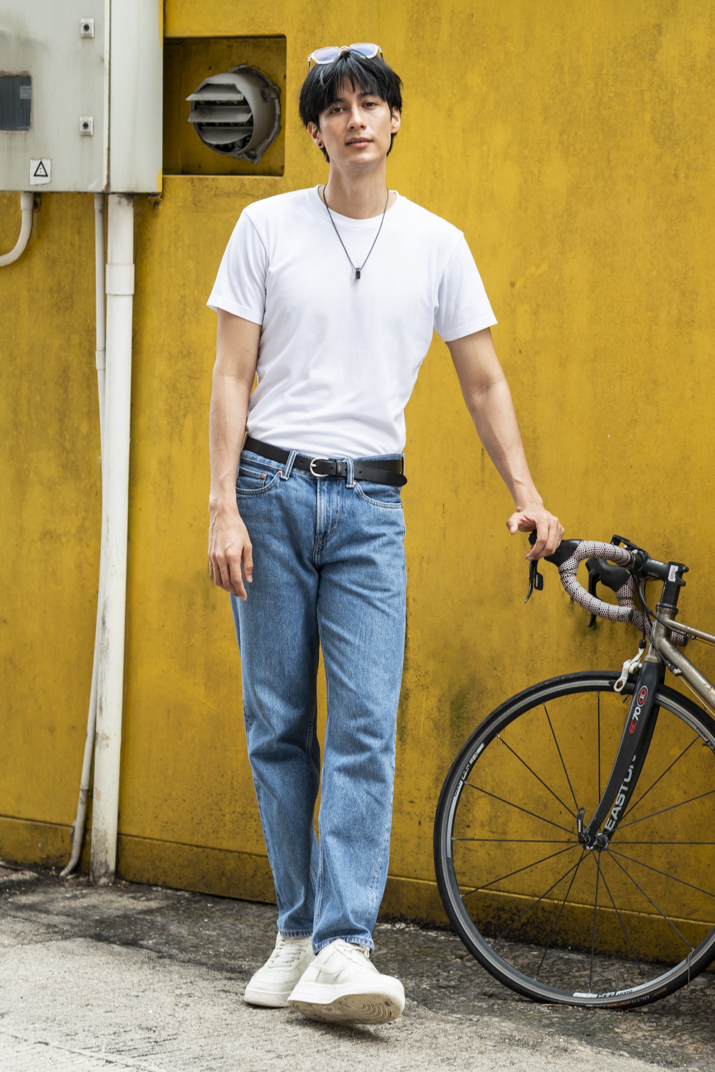 A young man with black hair, wearing a white T-shirt, blue jeans, and white sneakers, stands next to a bicycle with a yellow wall in the background. He has sunglasses on his head and is holding the bicycle with one hand.