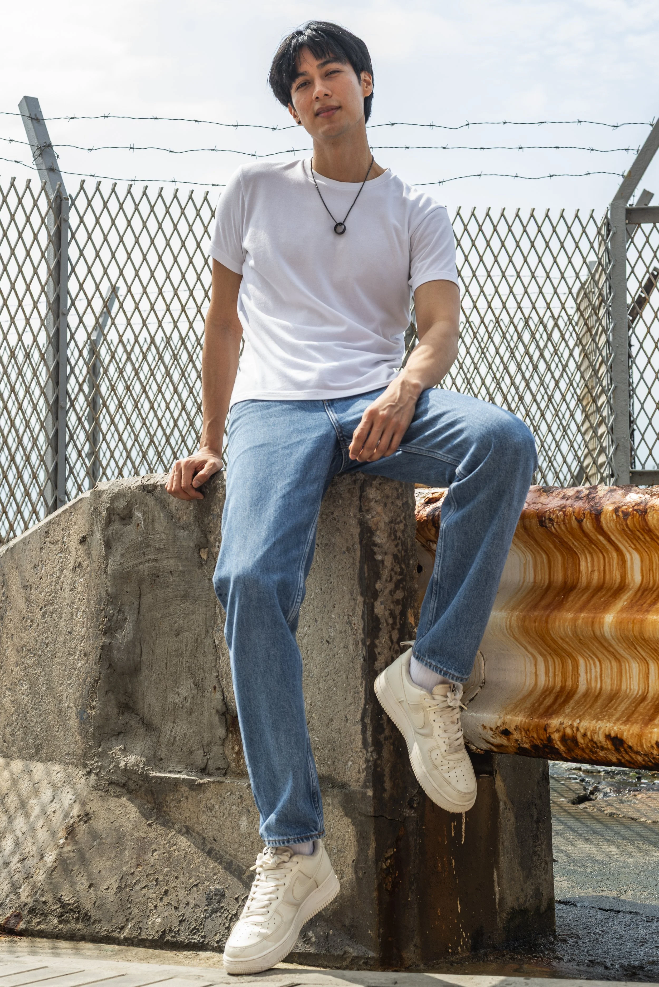 Young man wearing a white t-shirt, blue jeans, and white sneakers sitting on a large rock with a decorative stone structure, an industrial metal fence with barbed wire, and an overcast sky in the background.