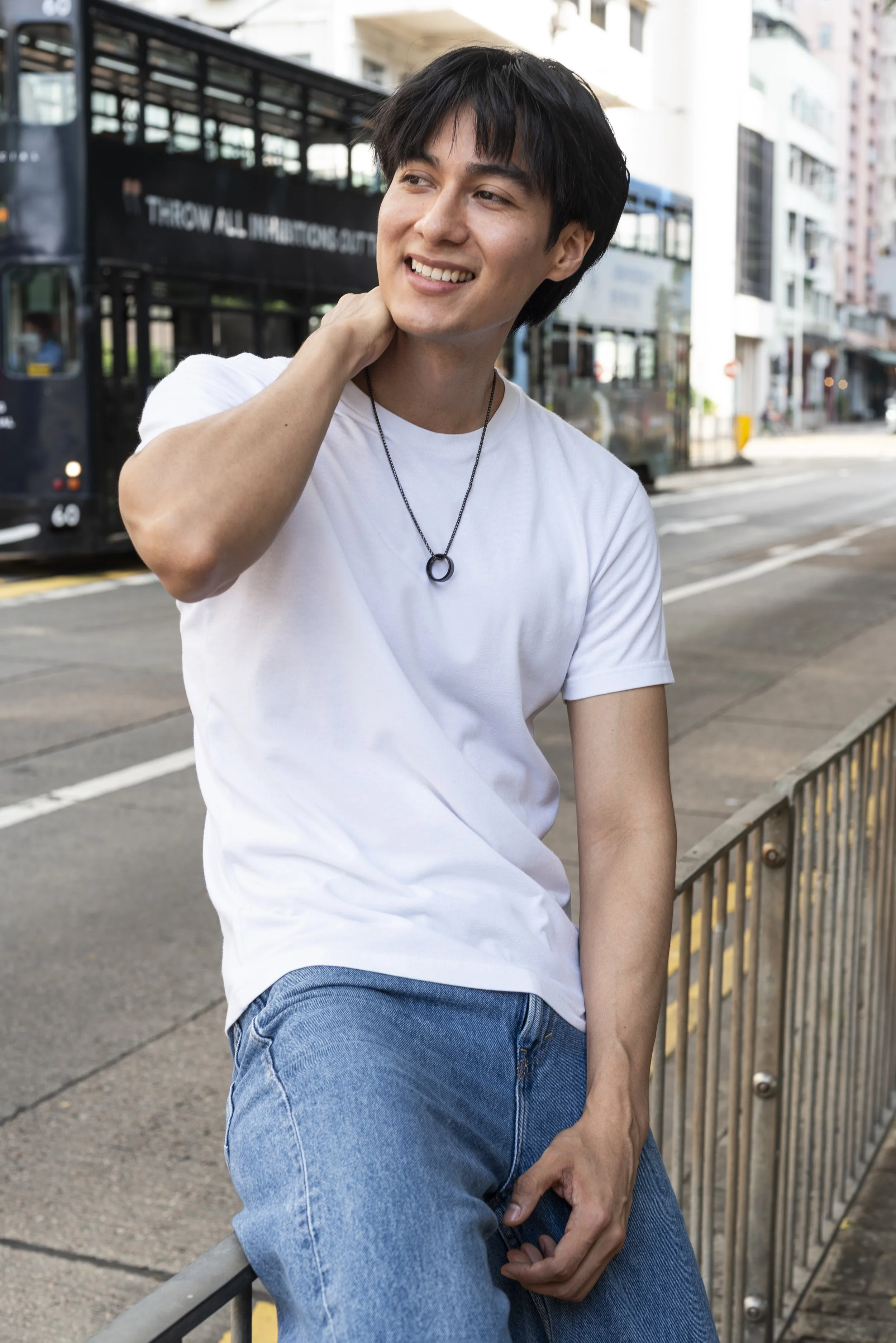 A young man with dark hair smiling and sitting on a city street fence, wearing a white T-shirt, blue jeans, and a necklace, with a city bus and tall buildings in the background.