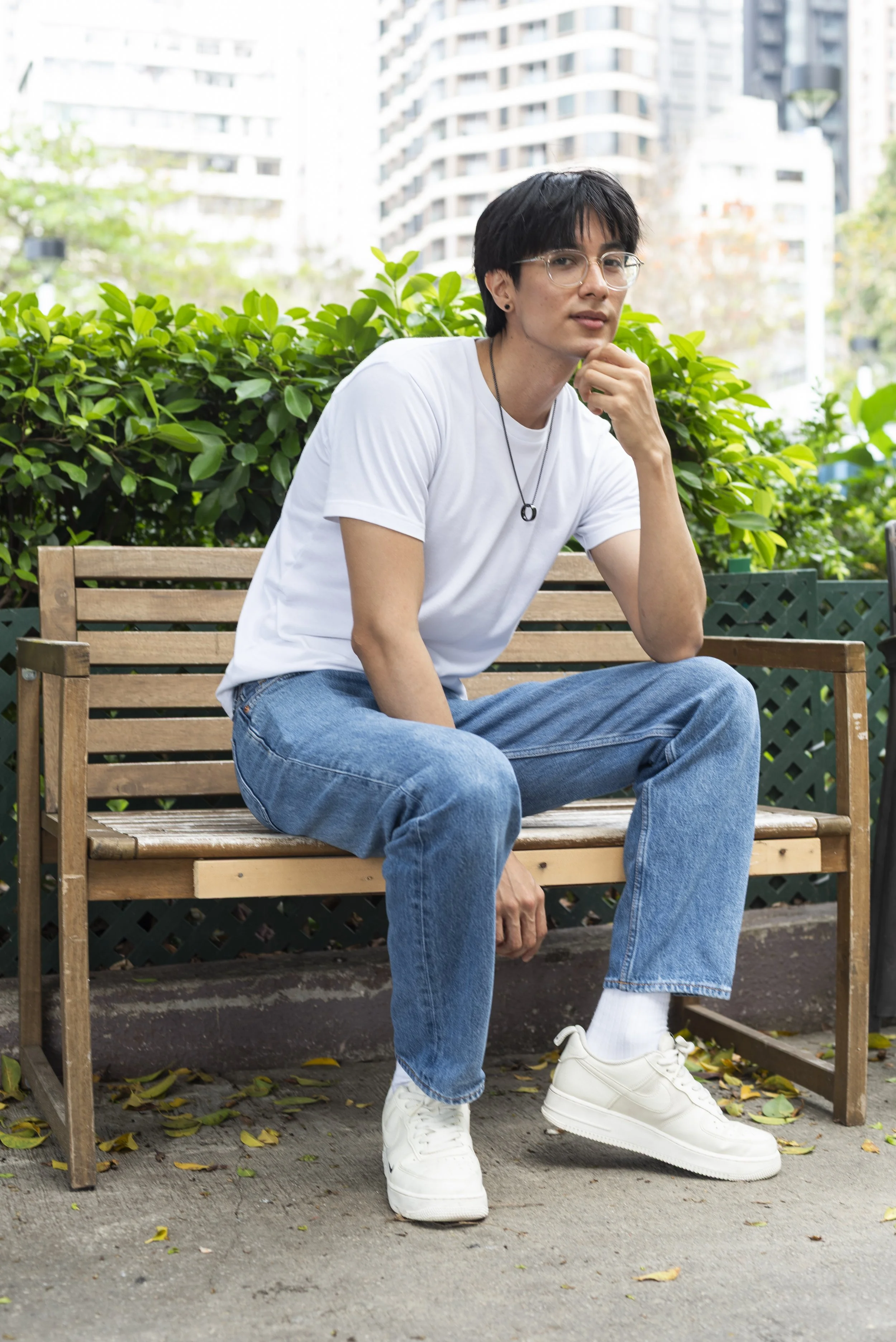 A person with short black hair, wearing glasses, a white t-shirt, blue jeans, and white sneakers, sitting on a wooden park bench with their right hand resting under their chin. The background shows tall buildings and green bushes, and there are falle