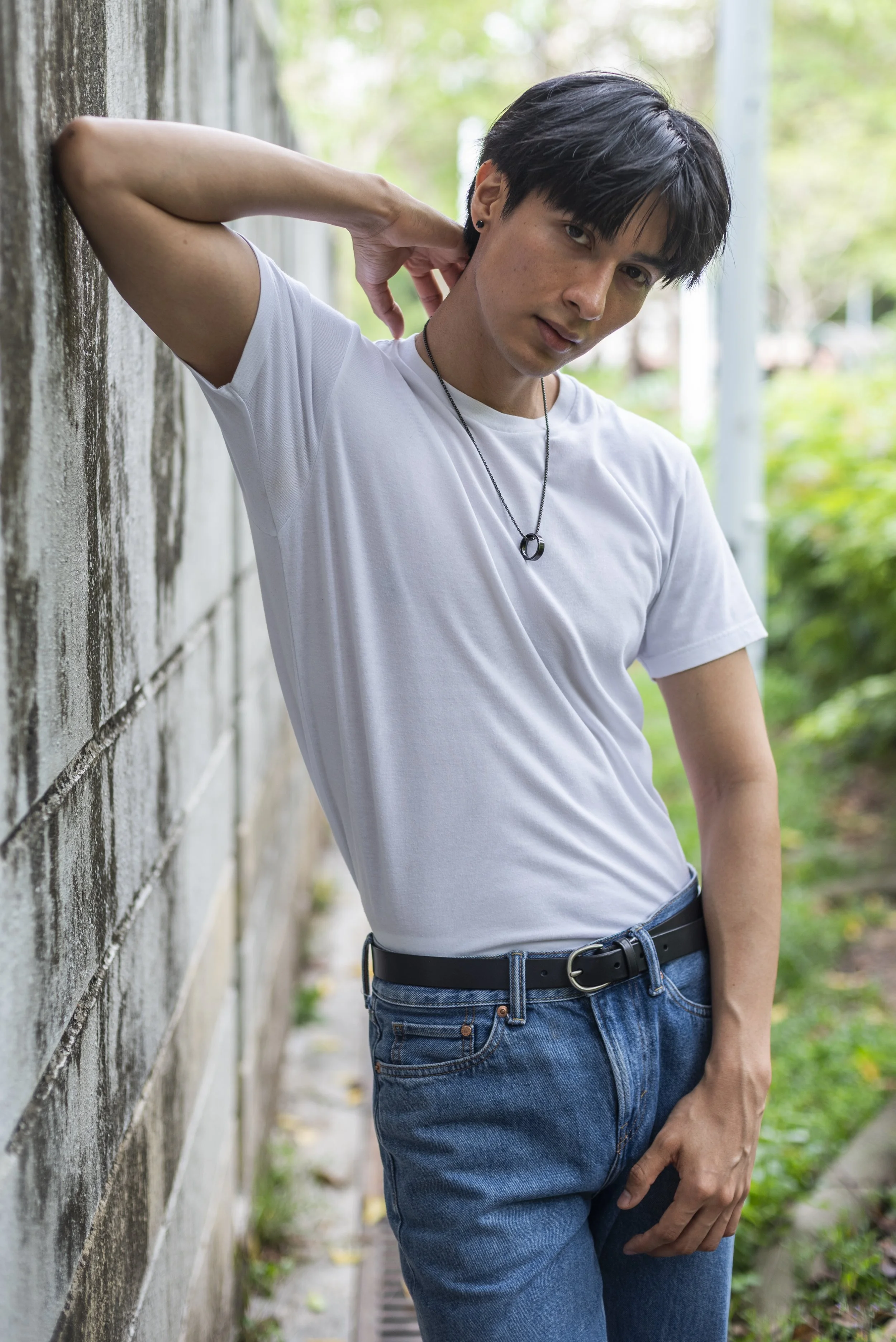 A young man with black hair, wearing a white t-shirt, blue jeans, and a black necklace, standing outdoors near a textured wall, touching his neck with one hand, and looking at the camera.
