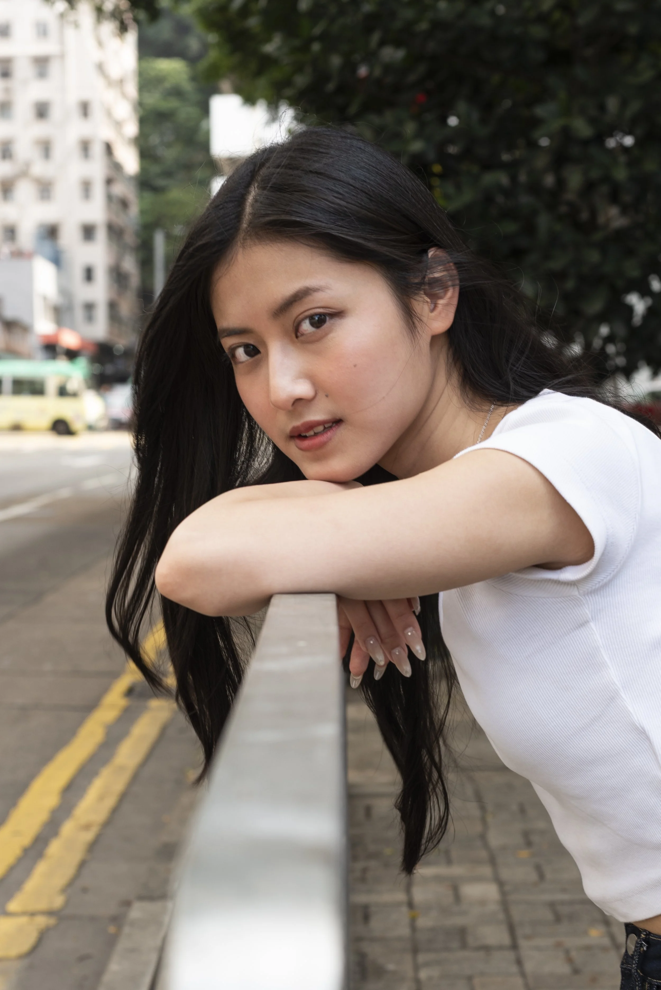 A young woman with long dark hair leaning on a railing outdoors in an urban setting, with buildings and trees in the background.