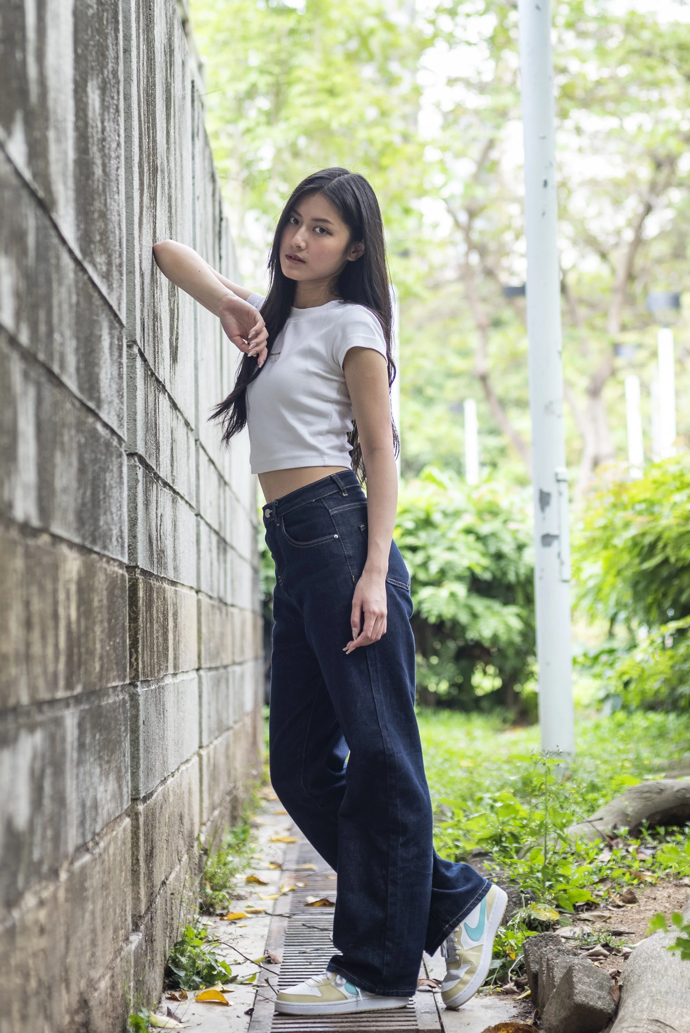 A young woman with long dark hair wearing a white crop top, dark jeans, and colorful Nike sneakers standing outdoors near a wooden fence and green trees, posing with one arm resting against the fence.