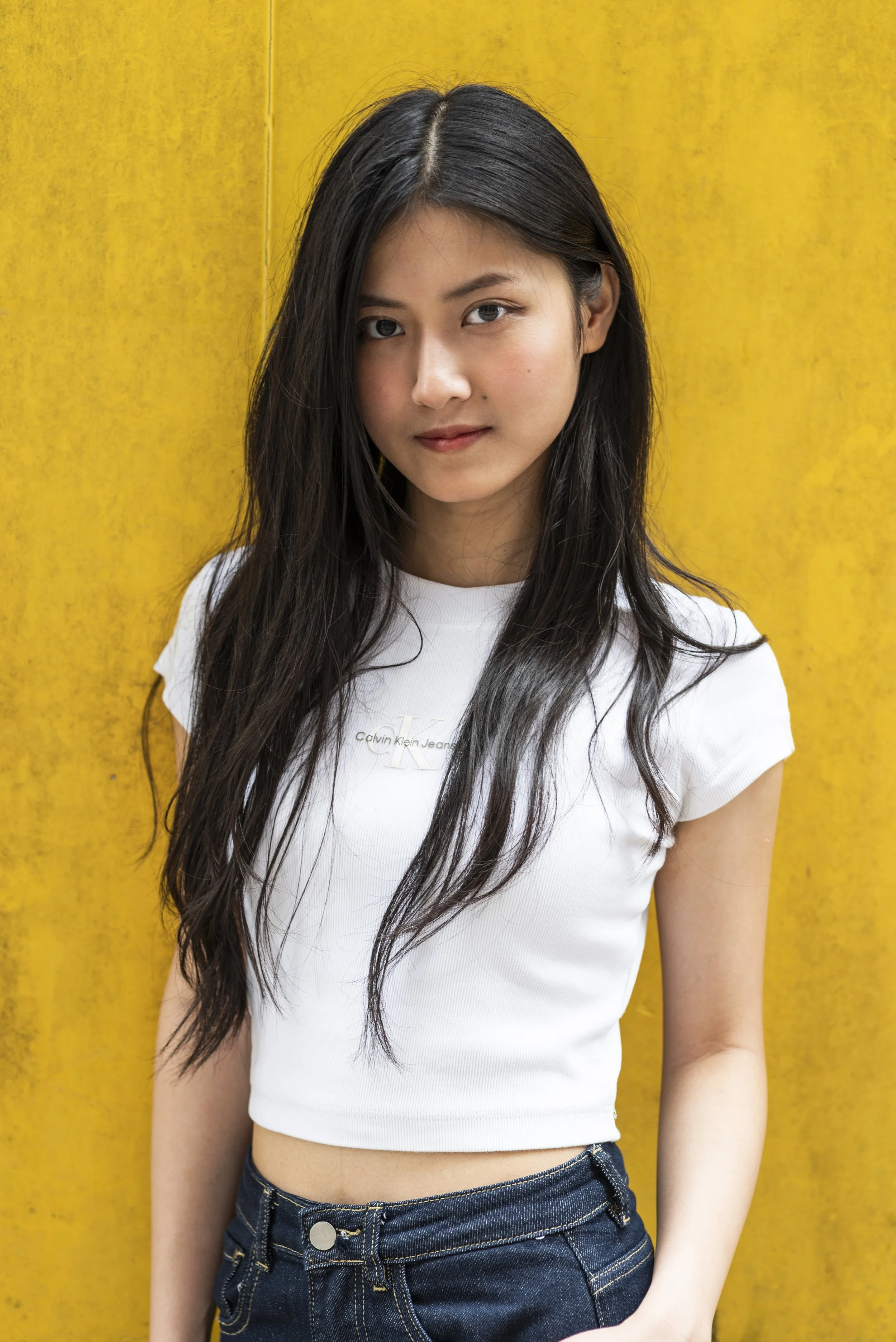 A young woman with long dark hair and light skin is standing against a yellow background, wearing a white Calvin Klein t-shirt and blue jeans.