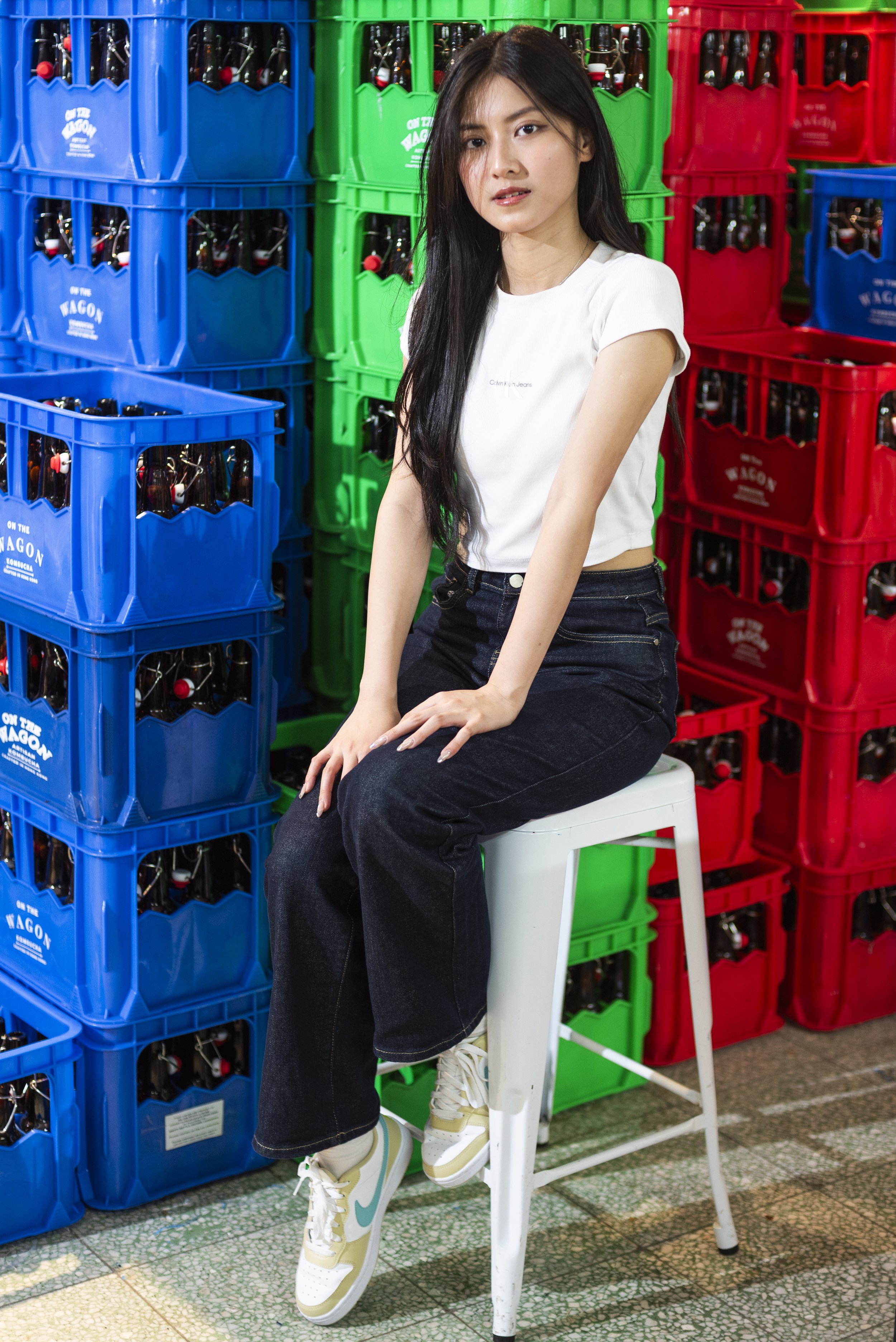 A young woman with long black hair is sitting on a white chair. She is wearing a white Calvin Klein t-shirt, black jeans, and white sneakers with lime green and light blue accents. She is surrounded by stacks of blue, green, and red plastic crates fi