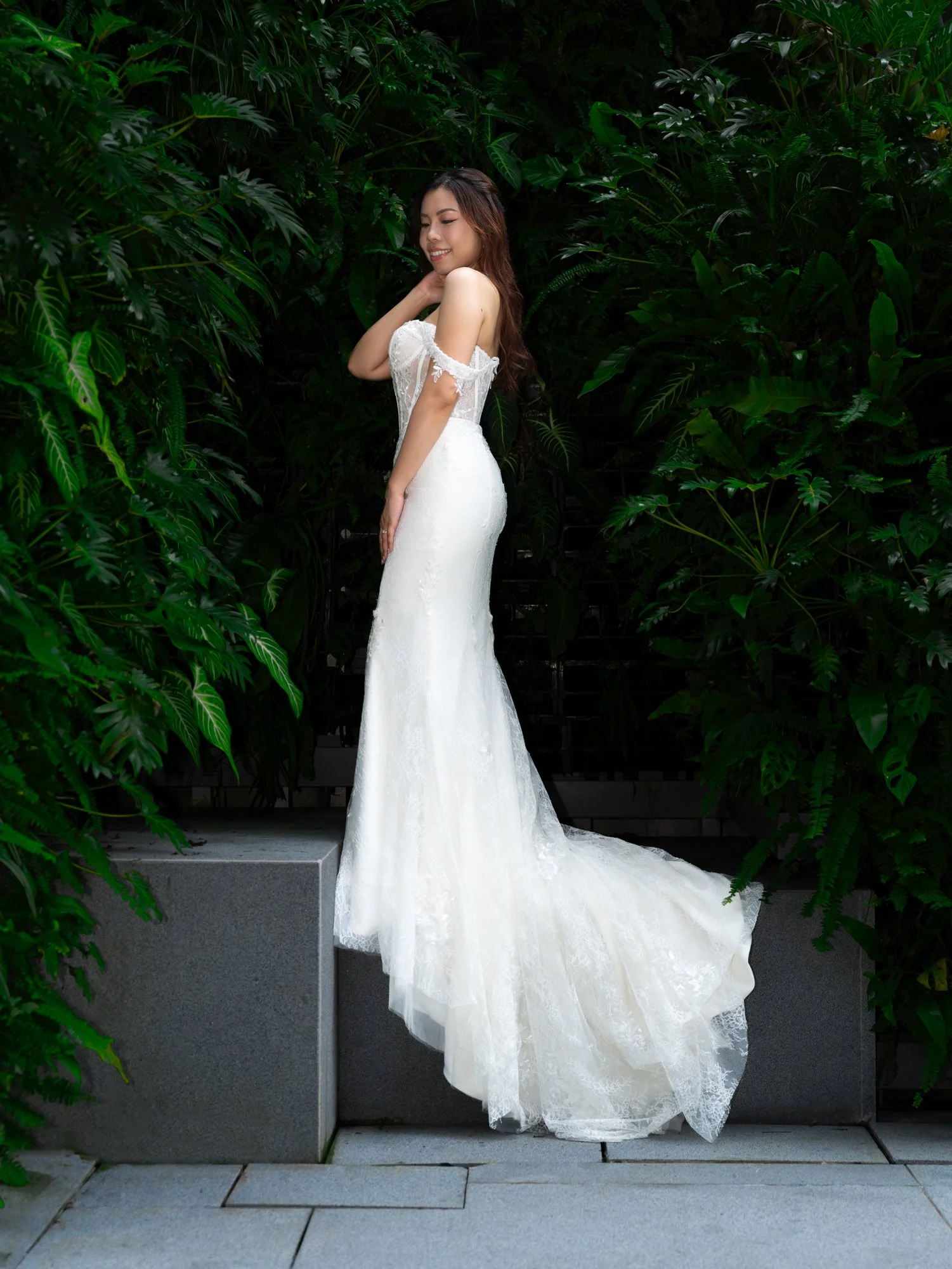 A woman in a white wedding dress standing next to lush green plants, with her hand touching her neck and smiling.