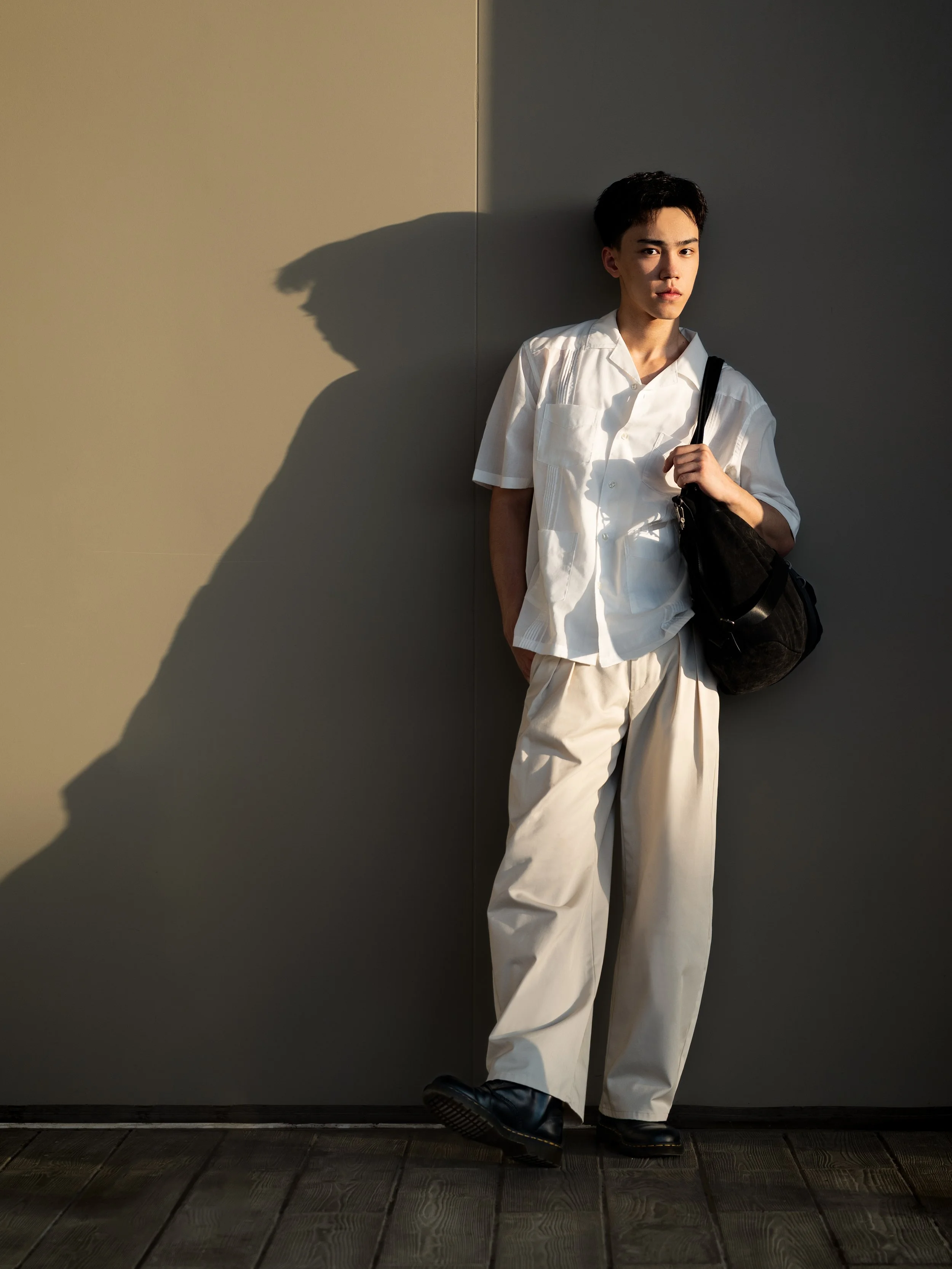Young man dressed in a white shirt and beige pants standing against a two-tone wall, carrying a black bag over his shoulder, with shadow cast on the wall.