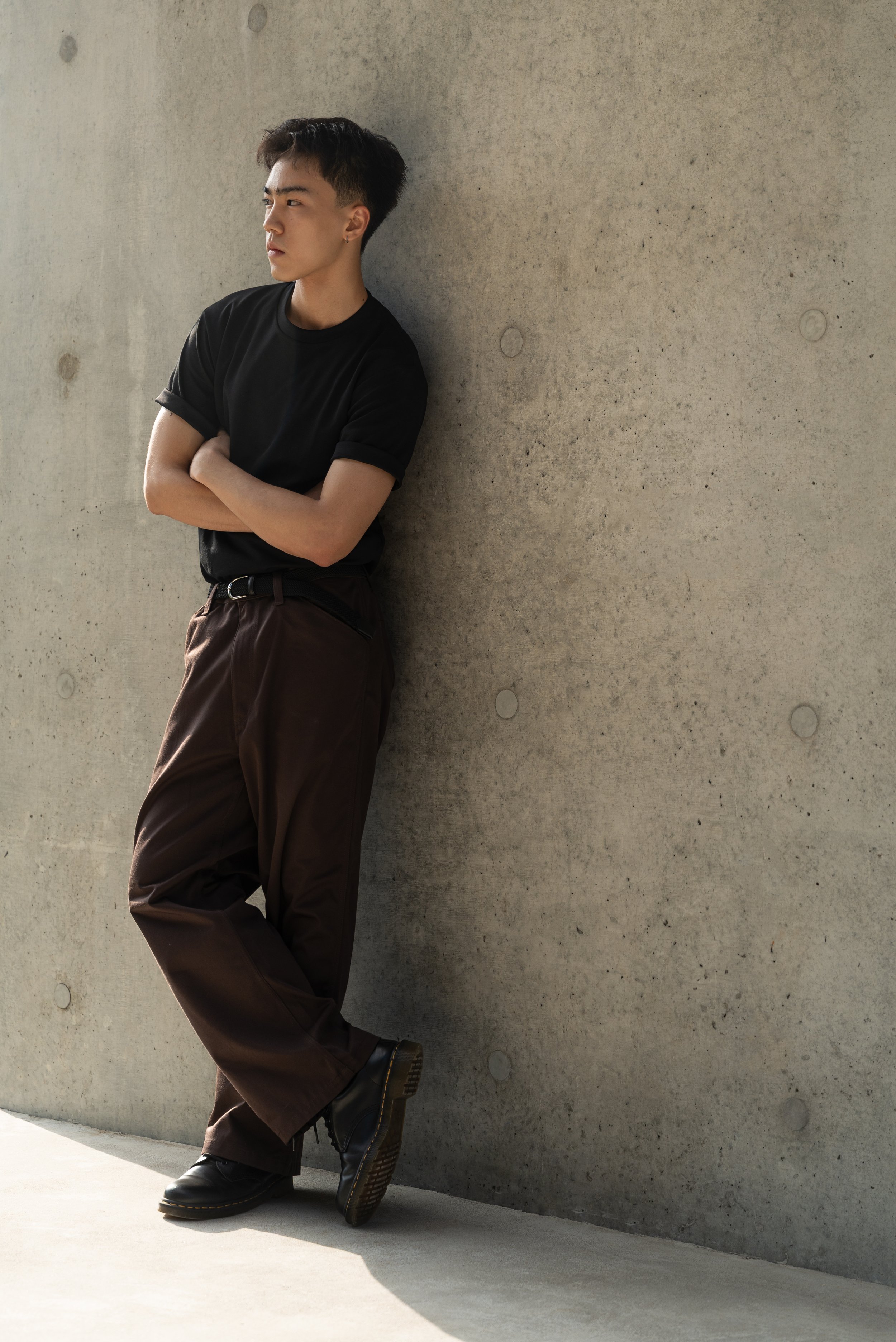 A young man leaning against a concrete wall with arms crossed, wearing a black t-shirt, brown pants, and black shoes, looking to the side.