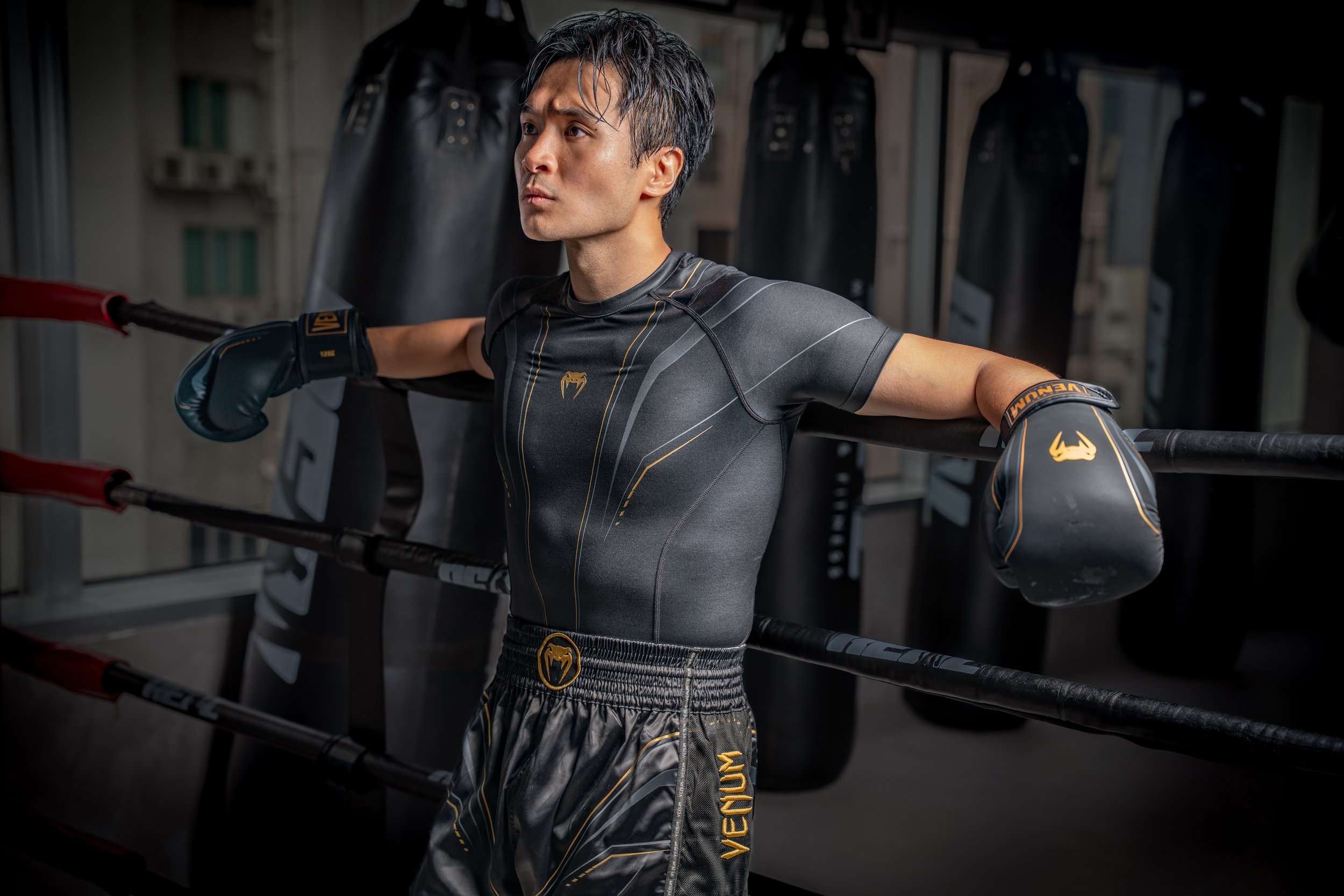 A man in a black and gold performance gear with boxing gloves, standing in a boxing gym, resting with his arms on boxing rings.