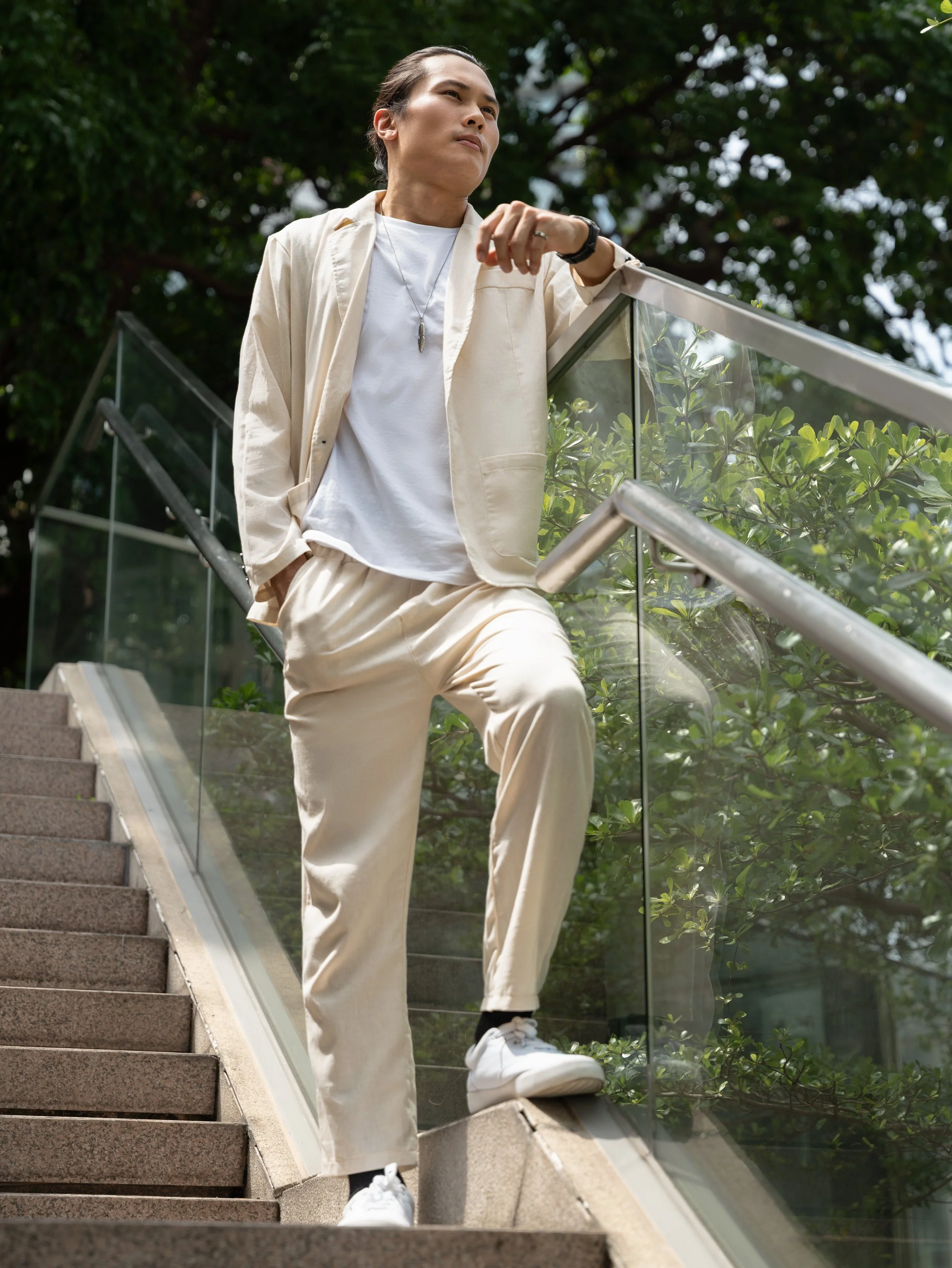Person leaning on a glass railing outdoors, wearing a white t-shirt, beige suit, white sneakers, and a necklace, with green trees in the background.