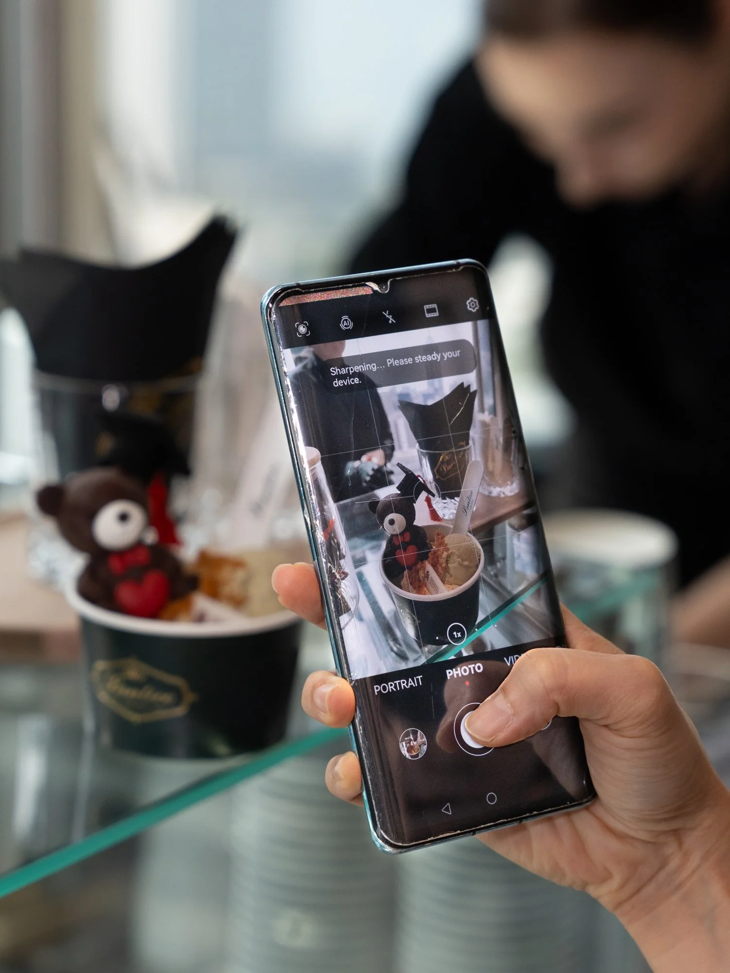 Person taking a photo of a cup with a stuffed reindeer figure using a smartphone. The phone screen displays the camera app in photo mode, with the message "Sharpening... Please steady your device."