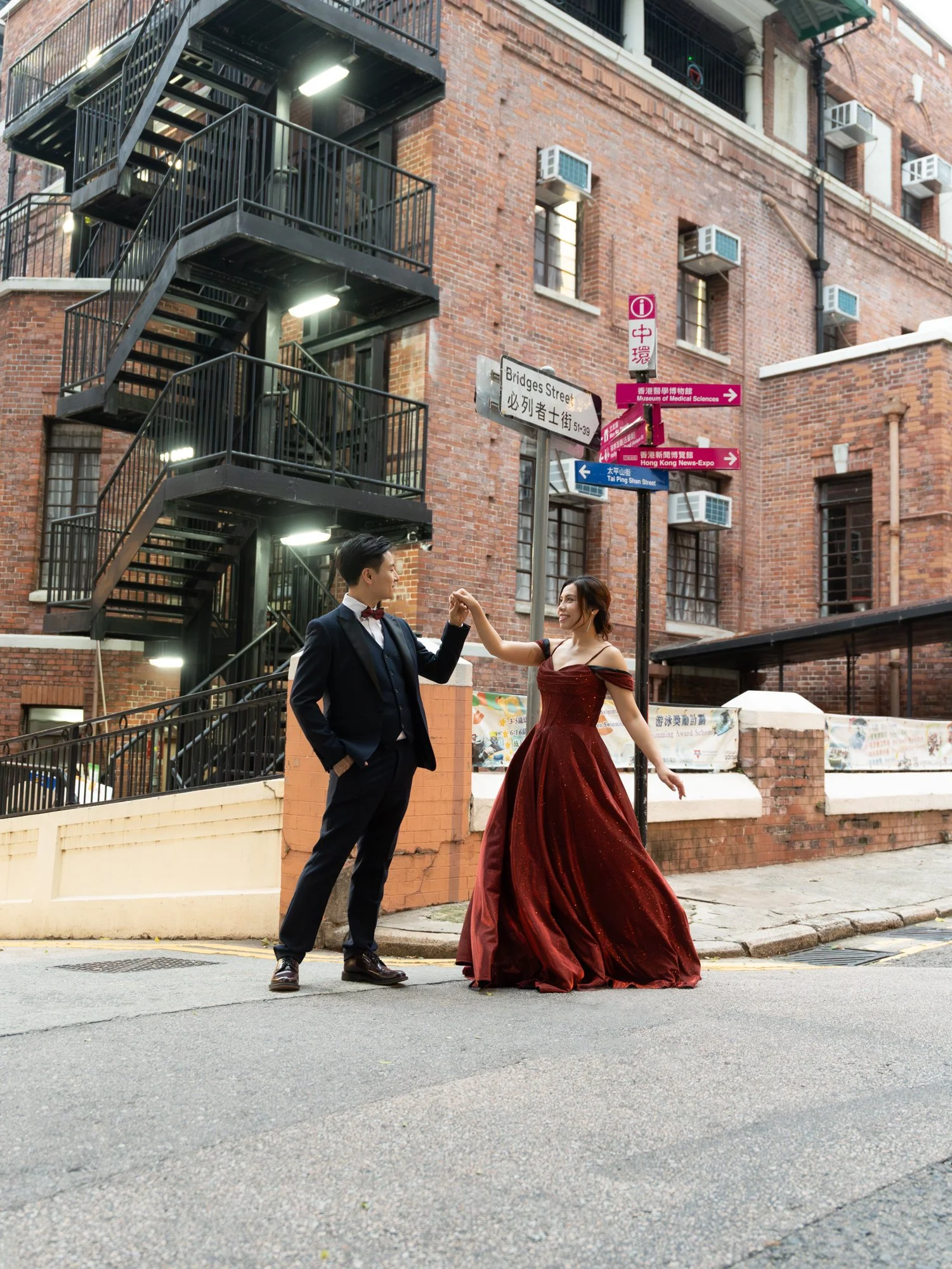 A couple dancing on a city street in front of brick buildings with fire escape stairs and street signs, one wearing a tuxedo and the other wearing a red gown.