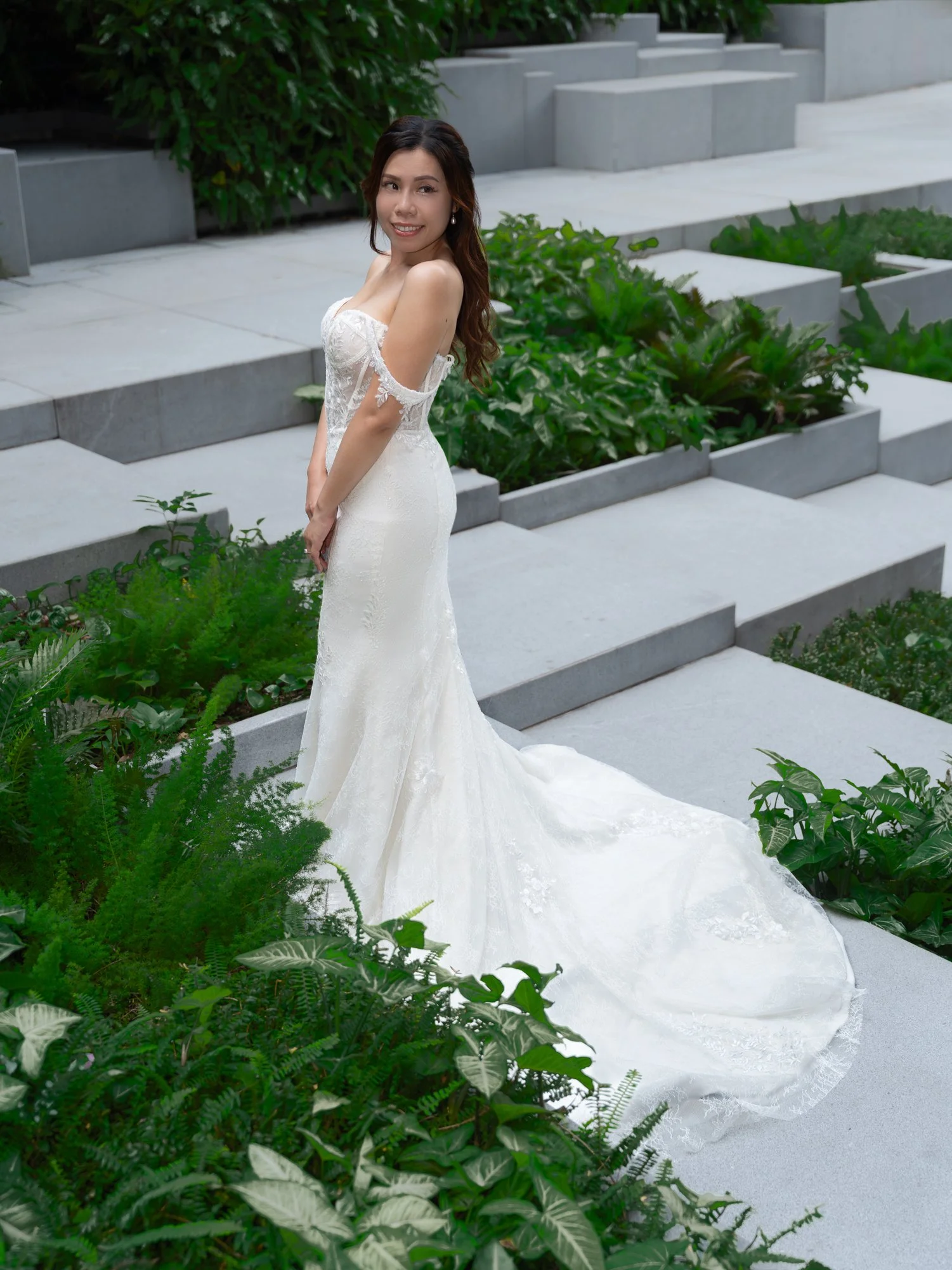 A woman in a white wedding dress standing outdoors on steps surrounded by lush green plants and foliage.