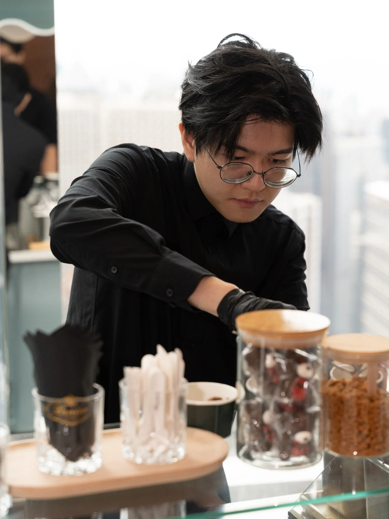 A person with dark hair and glasses wearing a black shirt, working behind a counter with jars and containers in a bright room with large windows.