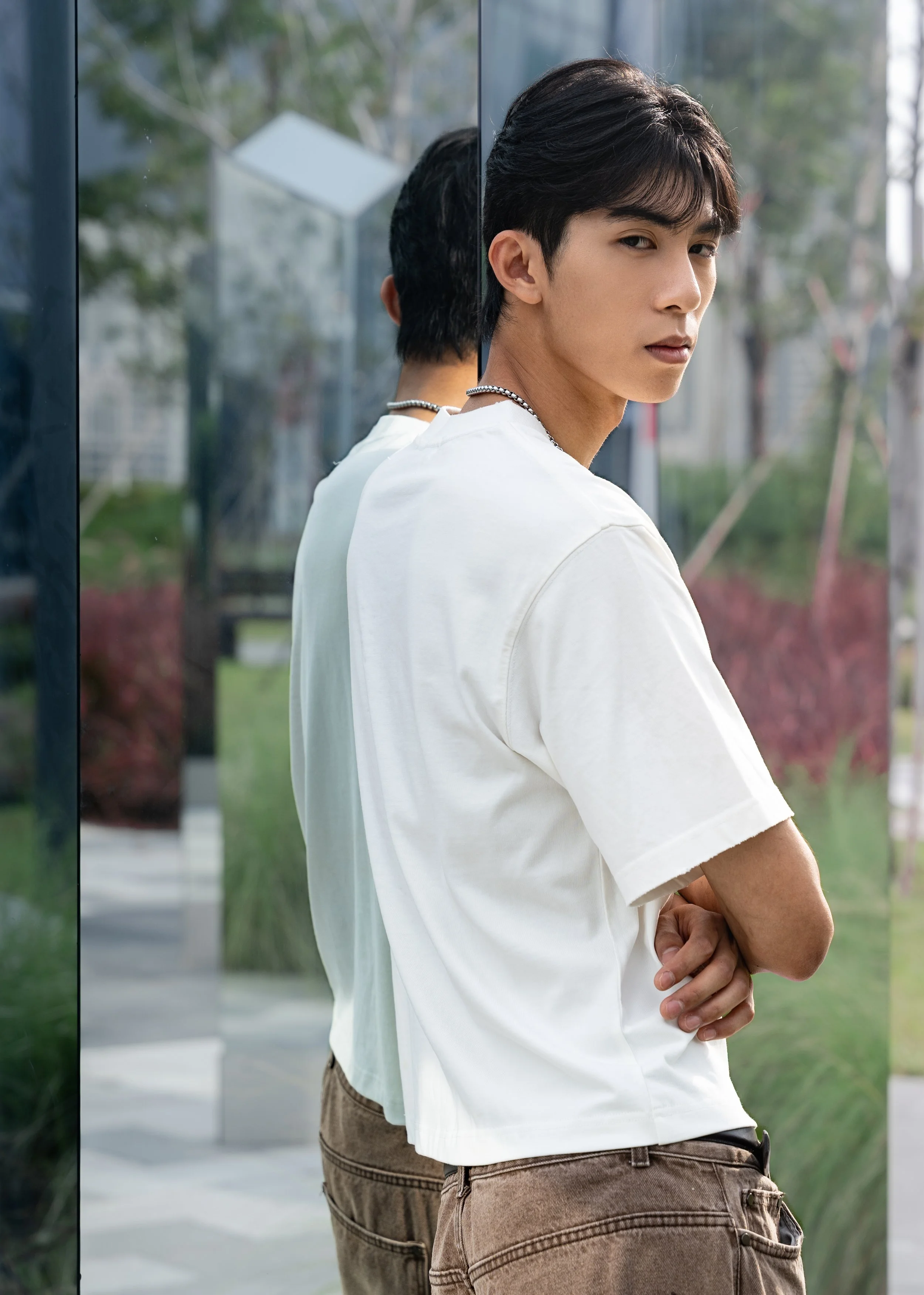 A young man with black hair and light skin stands outdoors, looking over his shoulder with a serious expression. He is wearing a white t-shirt and brown pants, and is positioned near a mirrored surface that reflects his back. The background features 