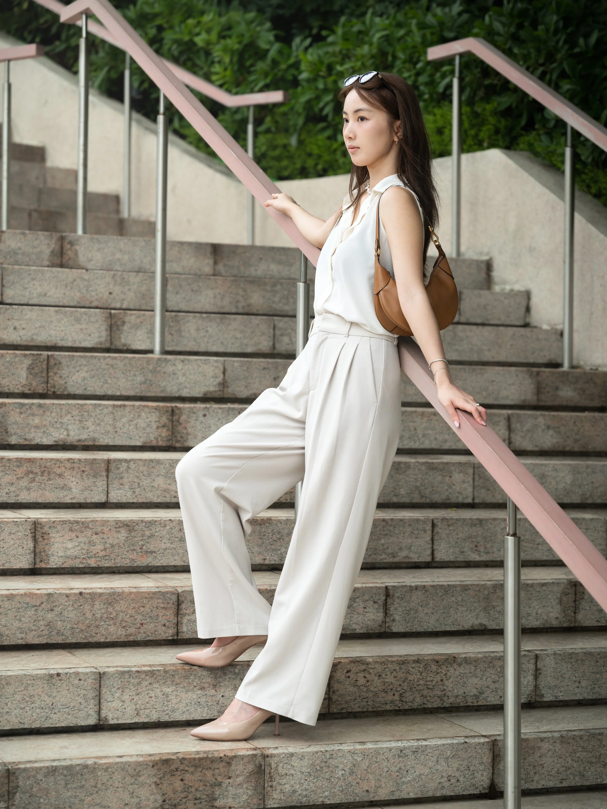 A young woman with long dark hair, wearing a white sleeveless blouse, cream-colored wide-legged trousers, nude heels, and sunglasses on her head, standing on outdoor stairs with a pink handrail and green foliage background.