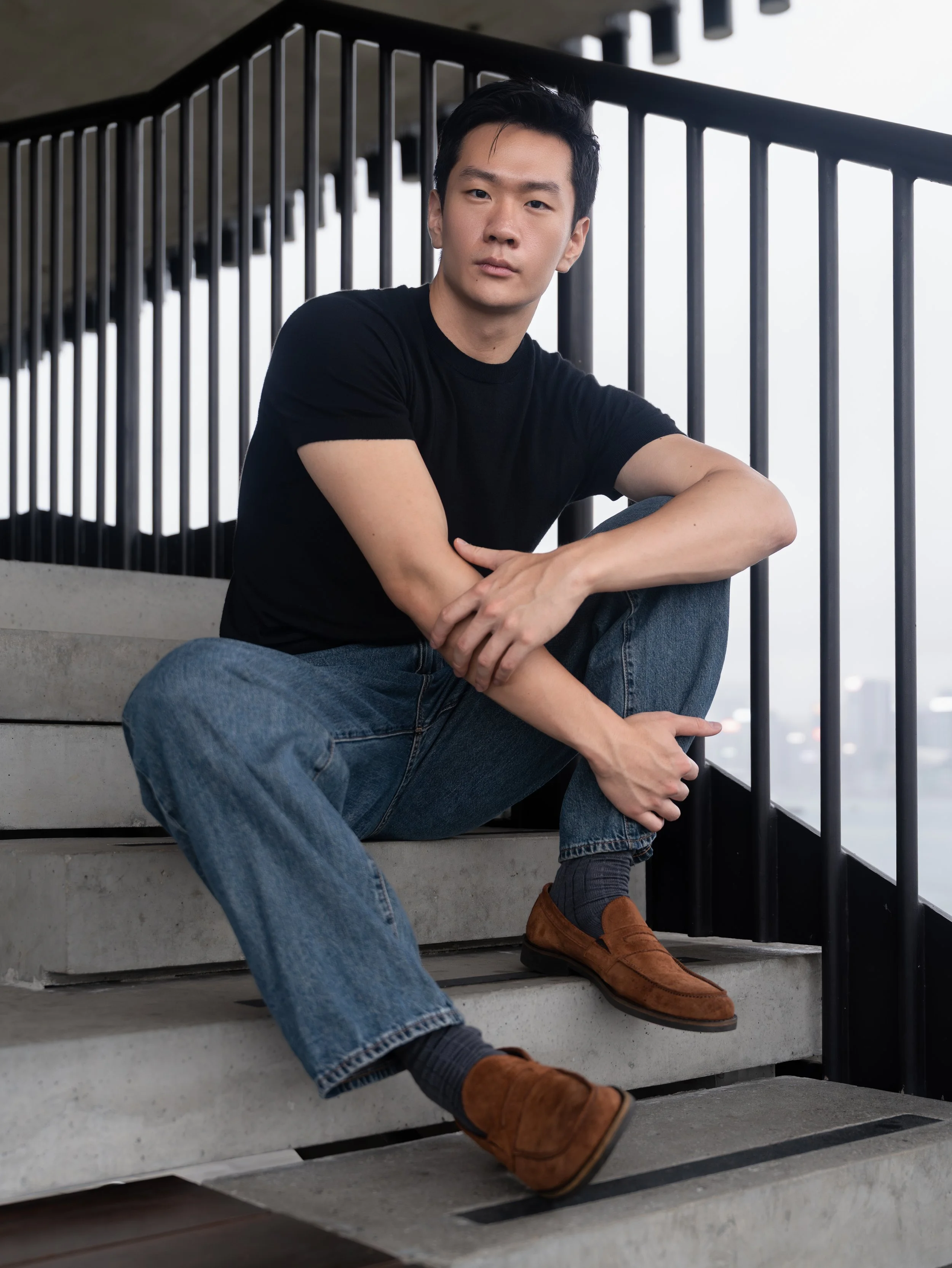 A young man in a black t-shirt and jeans sitting on concrete stairs near a black metal railing, with city buildings visible in the background.