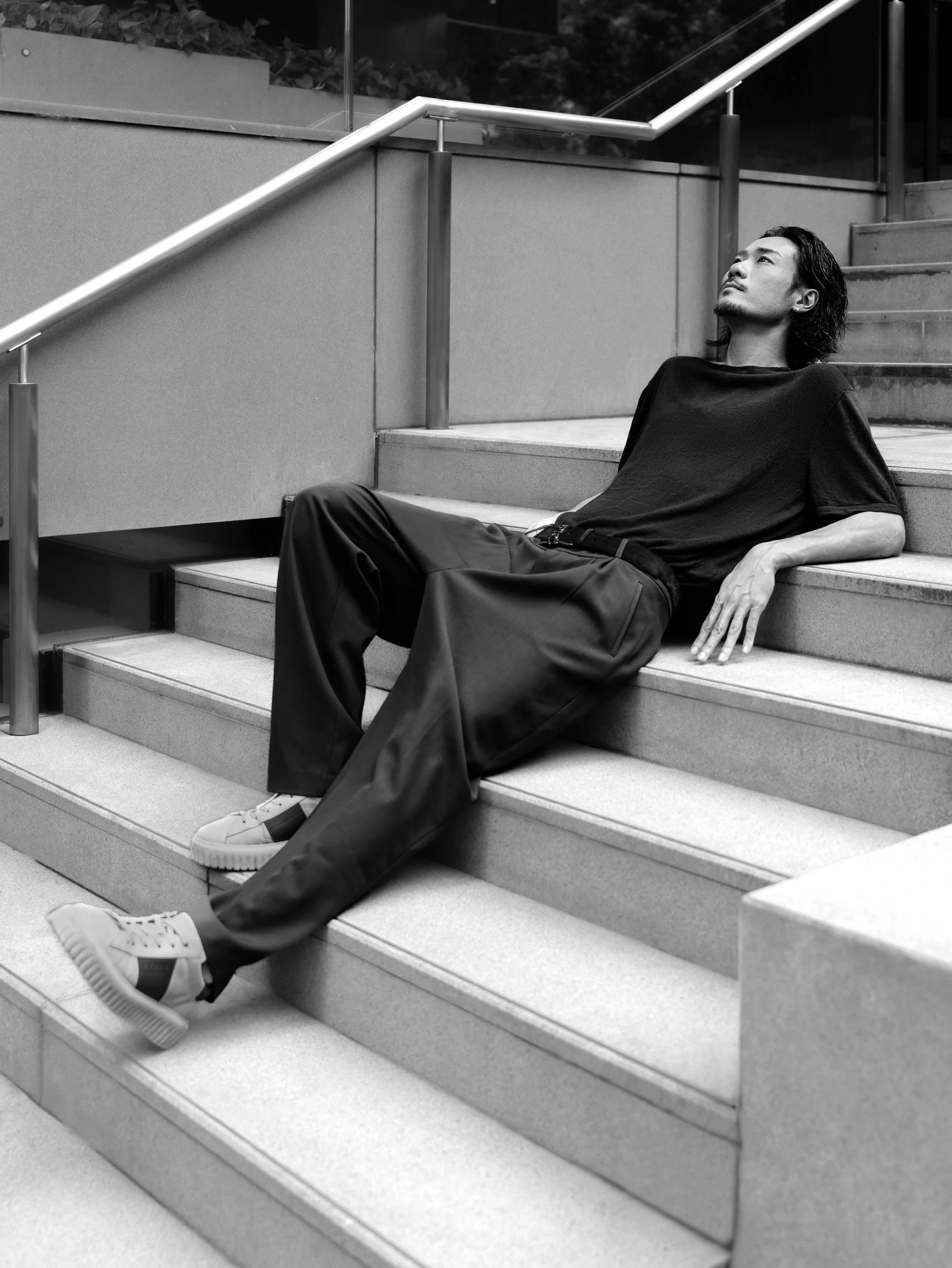 A man with long dark hair and a goatee relaxes on outdoor stairs, wearing a black t-shirt, dark pants, and white sneakers, looking upward.