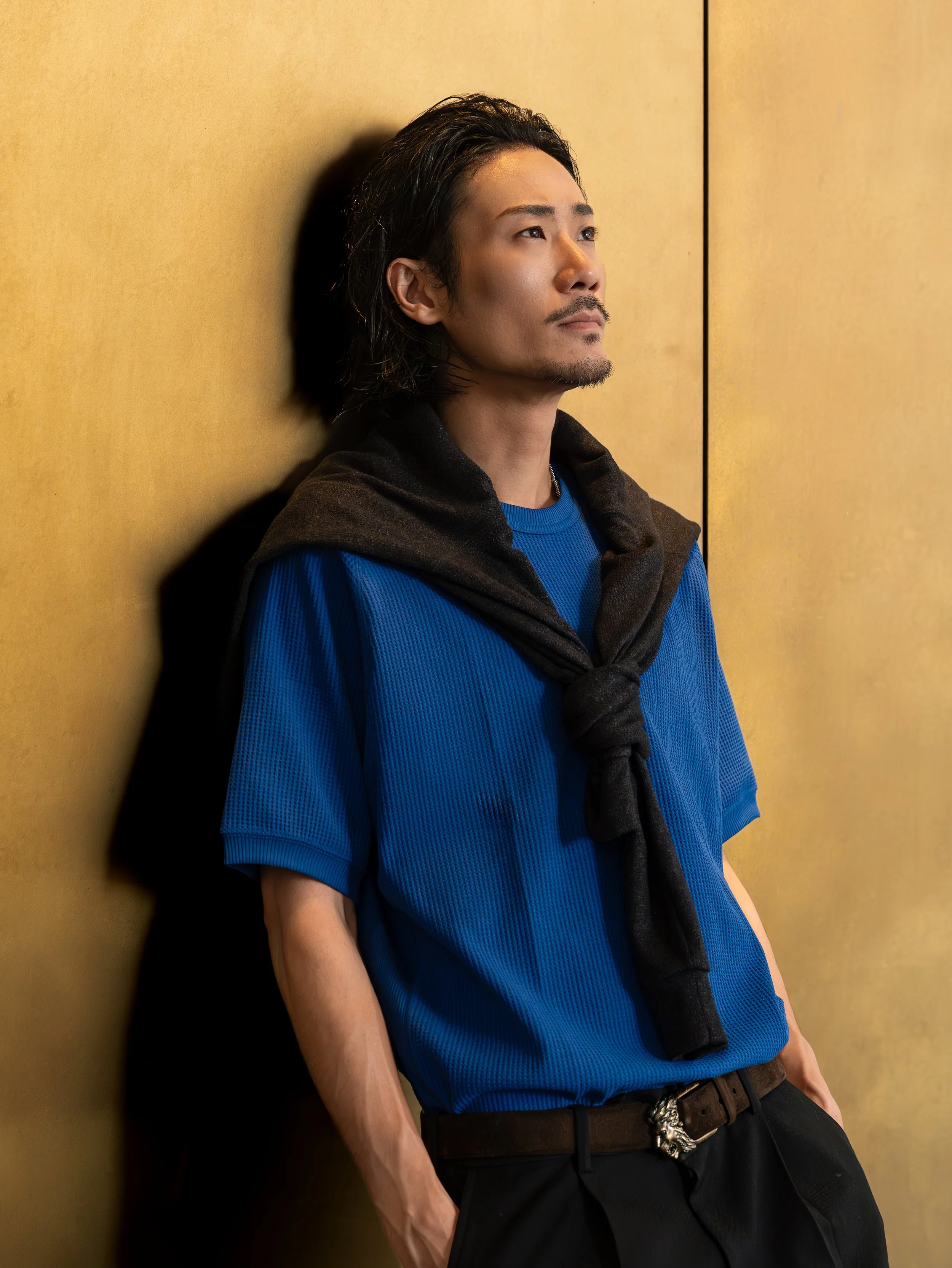 A man with long dark hair and a mustache is leaning against a gold wall, looking off into the distance. He is wearing a blue shirt with a black sweater draped over his shoulders, and black pants with a decorative tiger head belt buckle.